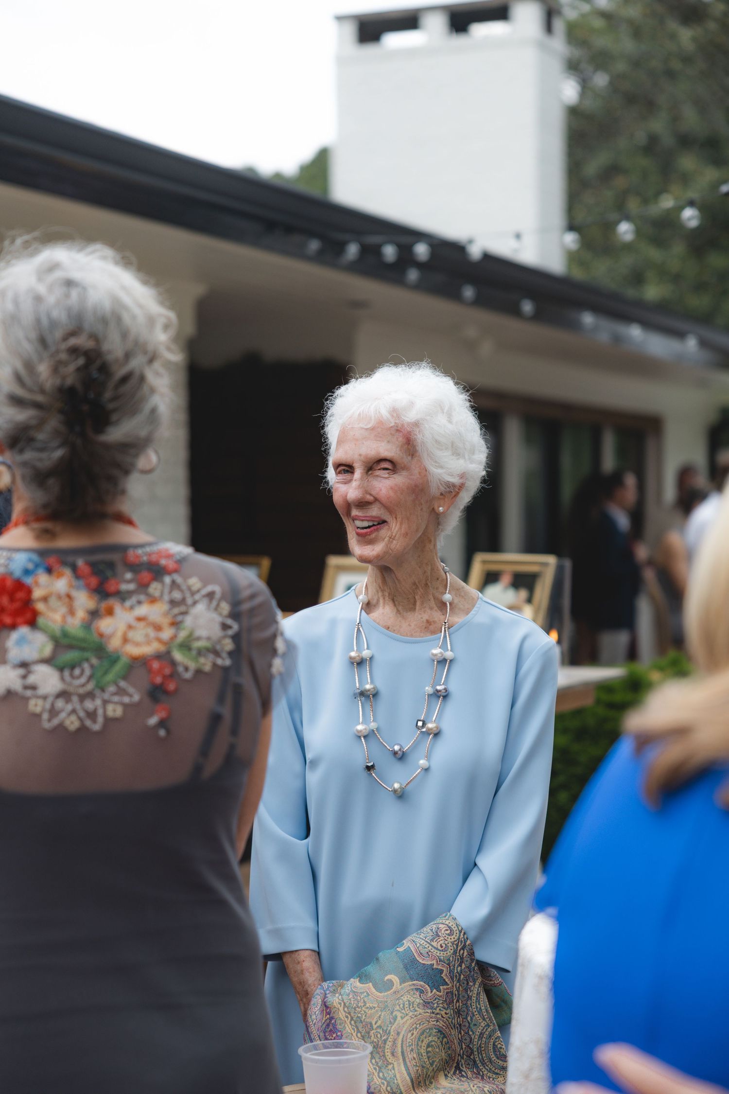 Elderly woman in light blue top and pearl necklace smiling at outdoor social gathering near a house.