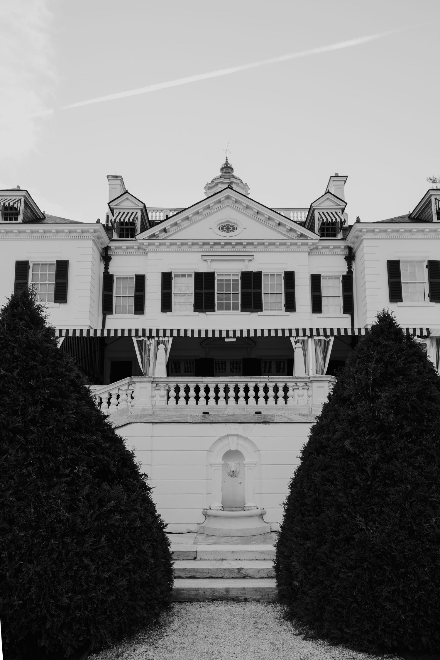 Black and white photo of an elegant colonial mansion with manicured hedges, balustrade staircase, and striped awnings.