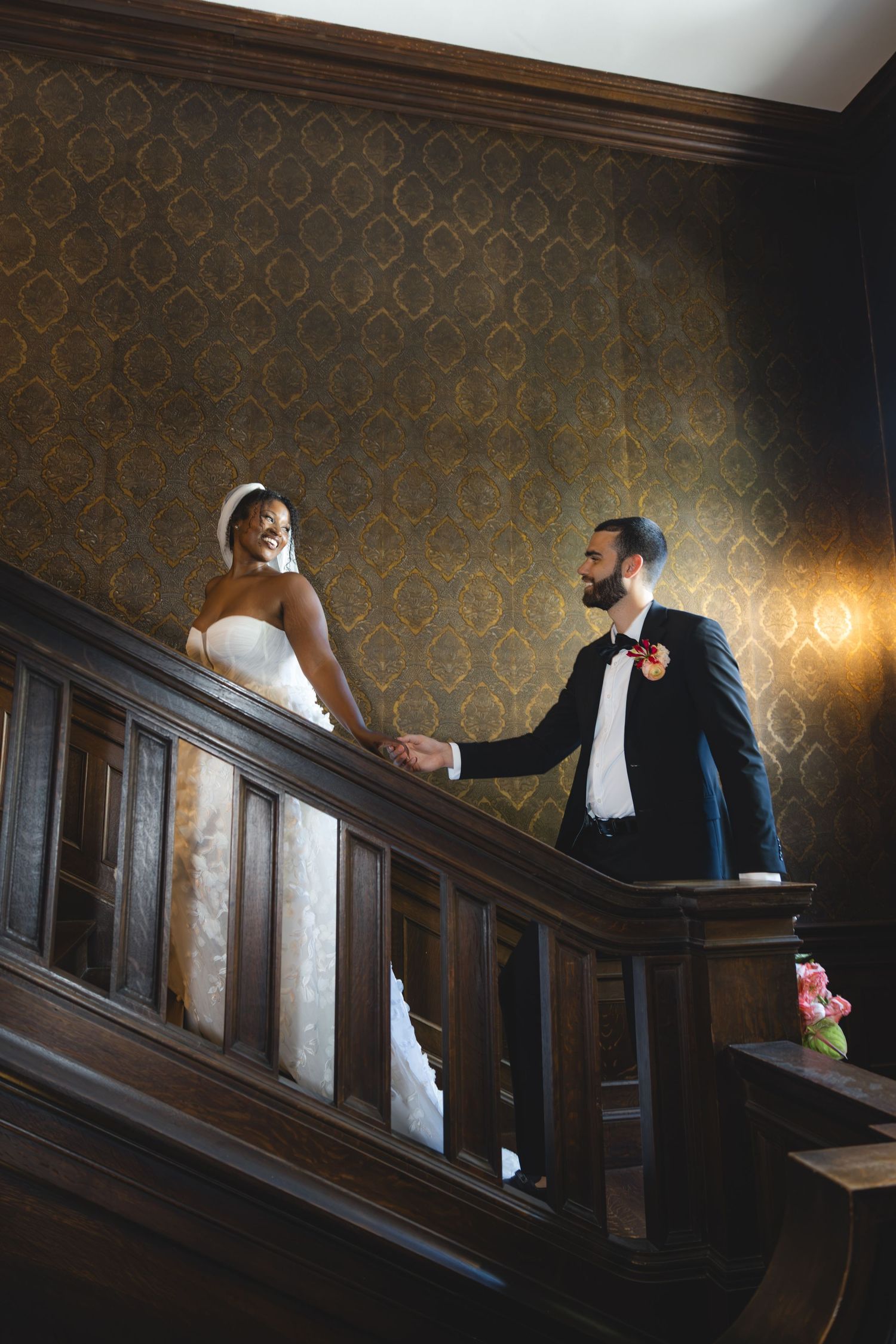 Bride and groom hold hands on elegant wooden staircase with ornate wallpaper in romantic wedding venue.