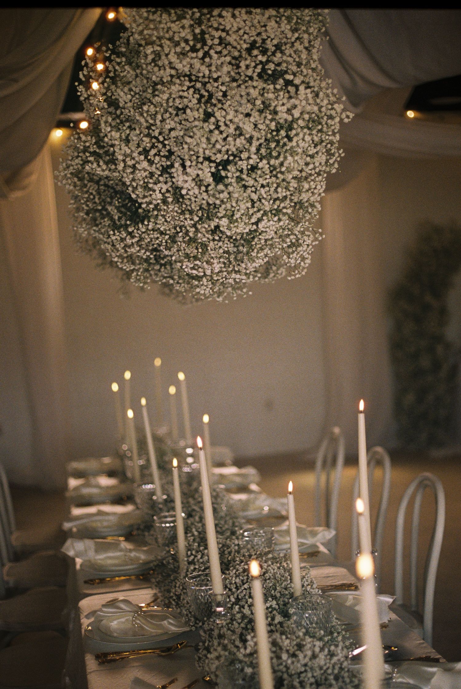 Elegant wedding reception table with white taper candles, baby's breath centerpieces, and a large floral chandelier overhead.