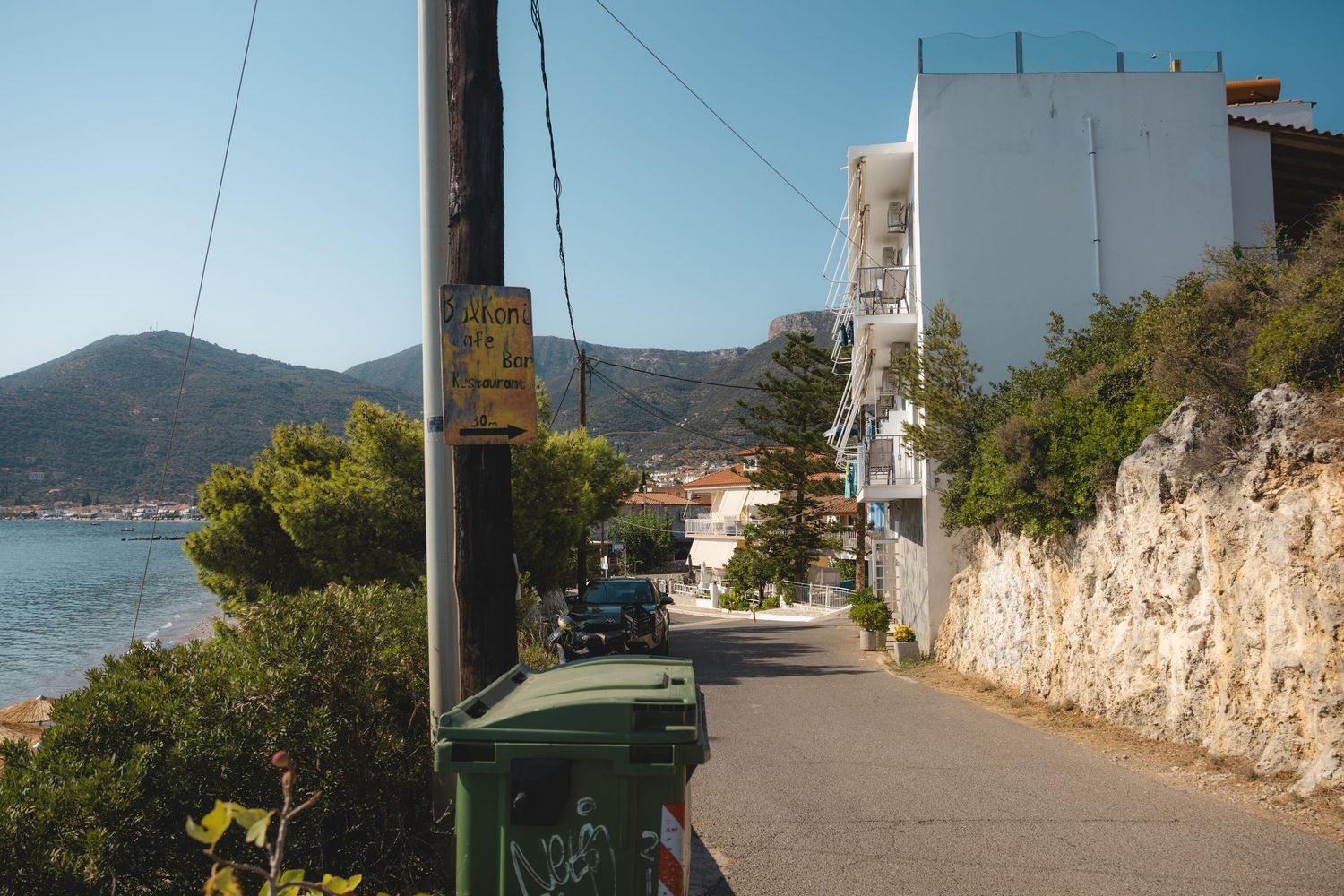 Narrow coastal road in Greek village with stone wall, white building, green bins, and sea views under clear blue sky.