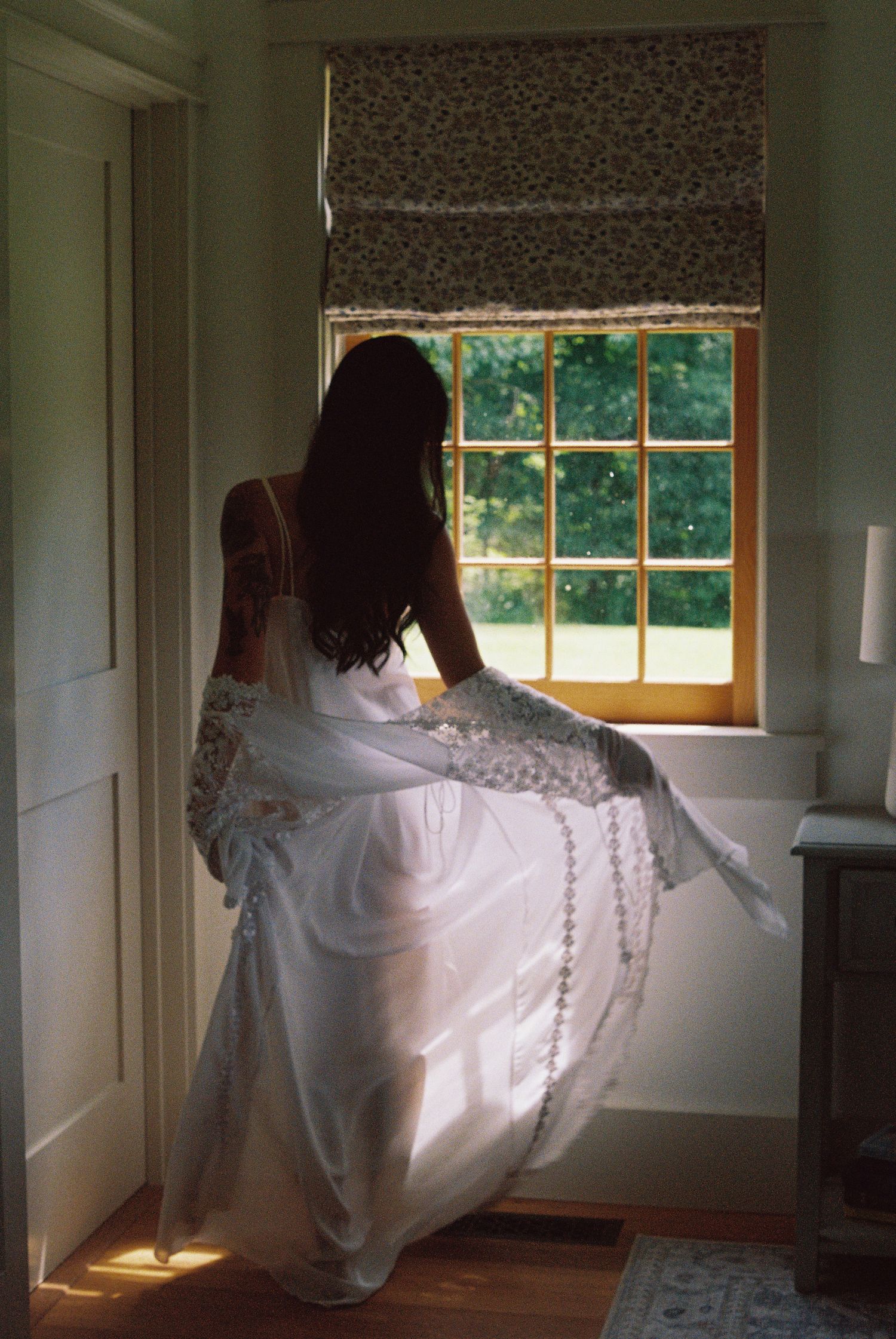 Bride in white wedding dress standing by sunlit window with floral roman shade, viewed from behind.