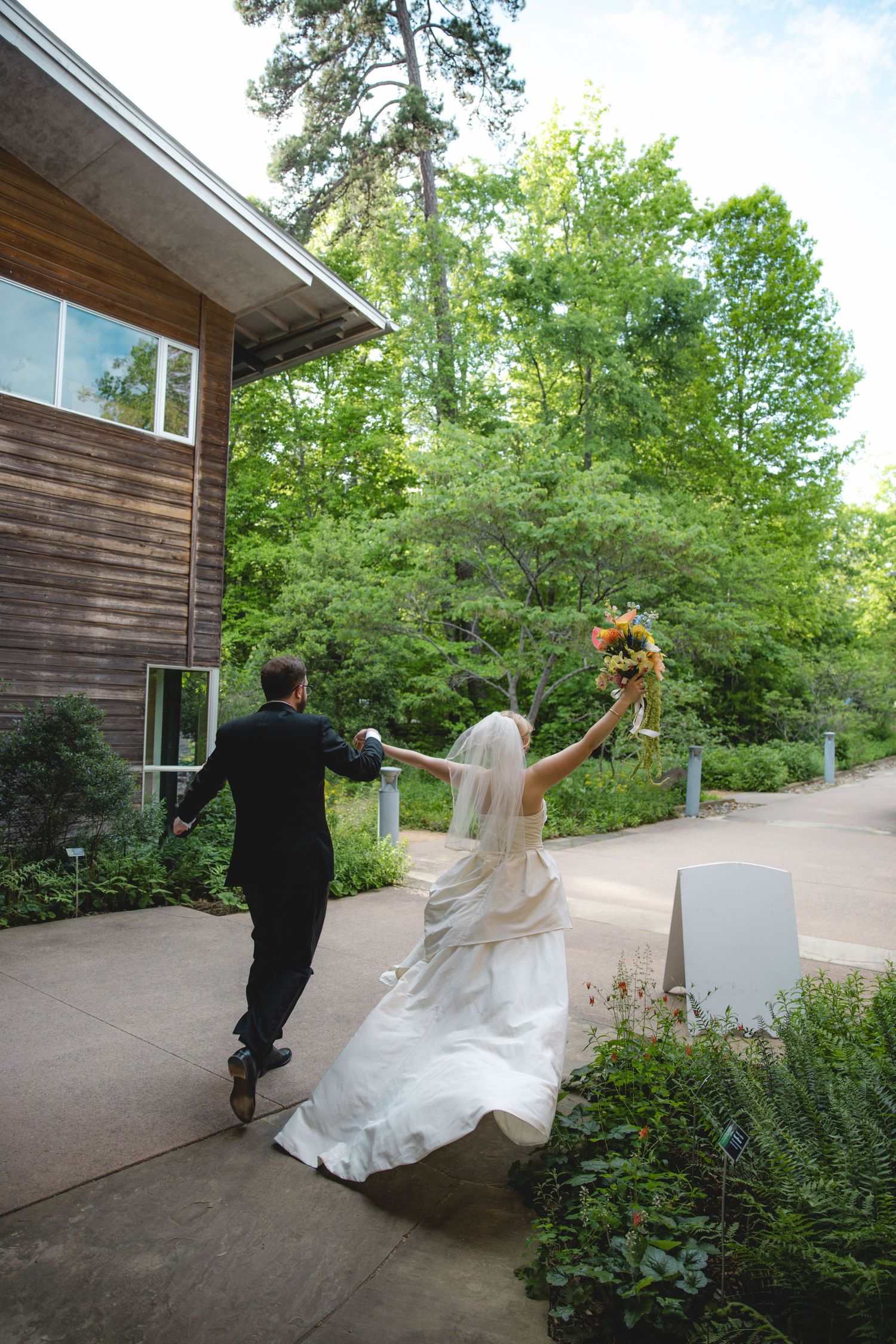 Bride and groom holding hands walking into venue, bride raising colorful bouquet, surrounded by lush green trees.