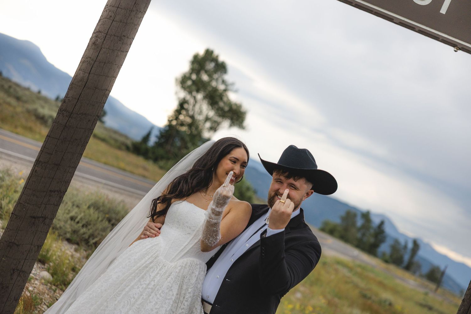 Bride and groom laughing while eating snacks outdoors with mountain lake scenery, bride in white gown, groom in black suit and cowboy hat.
