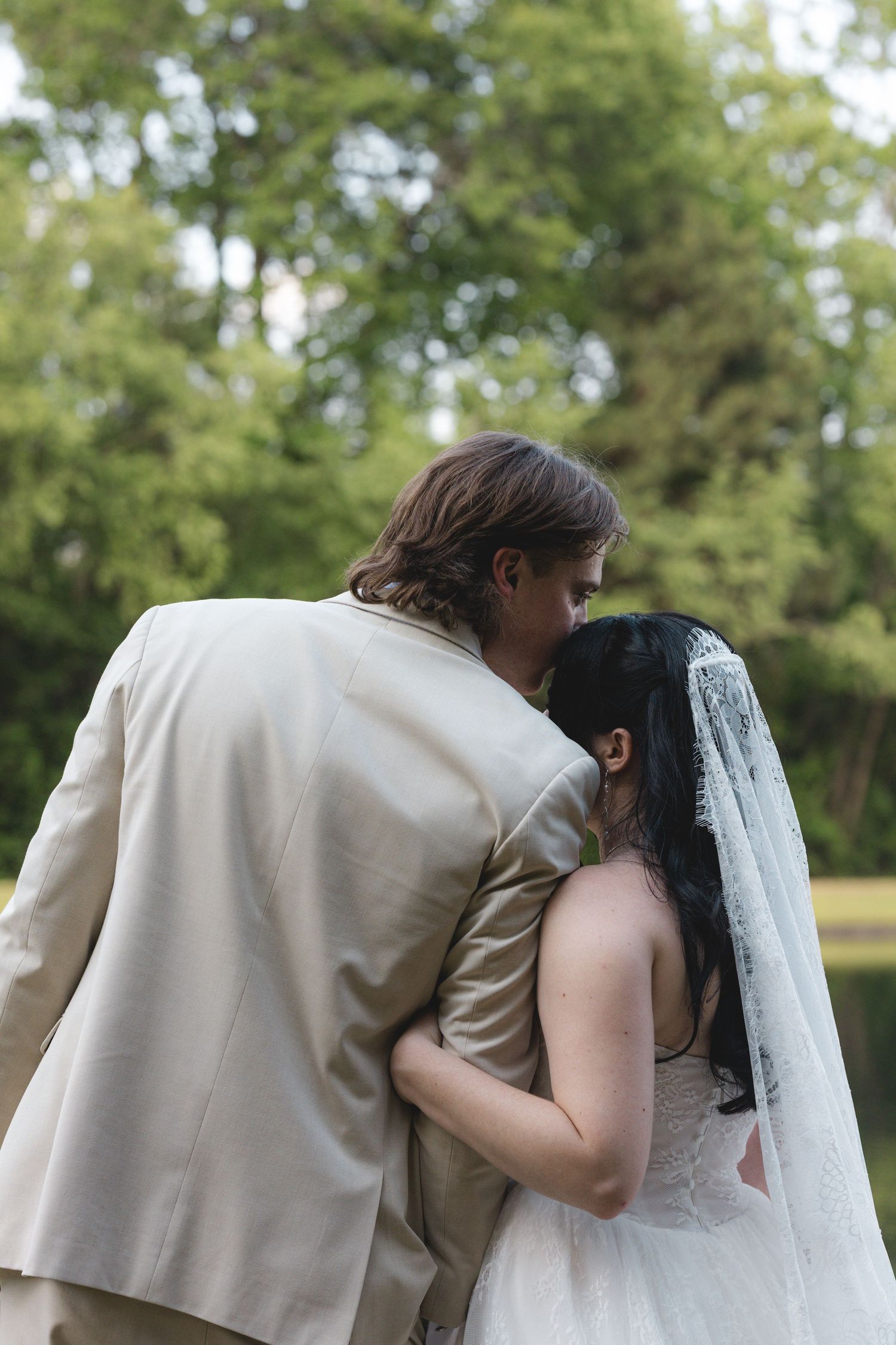 Bride and groom embracing from behind in outdoor wedding setting, surrounded by lush green trees.