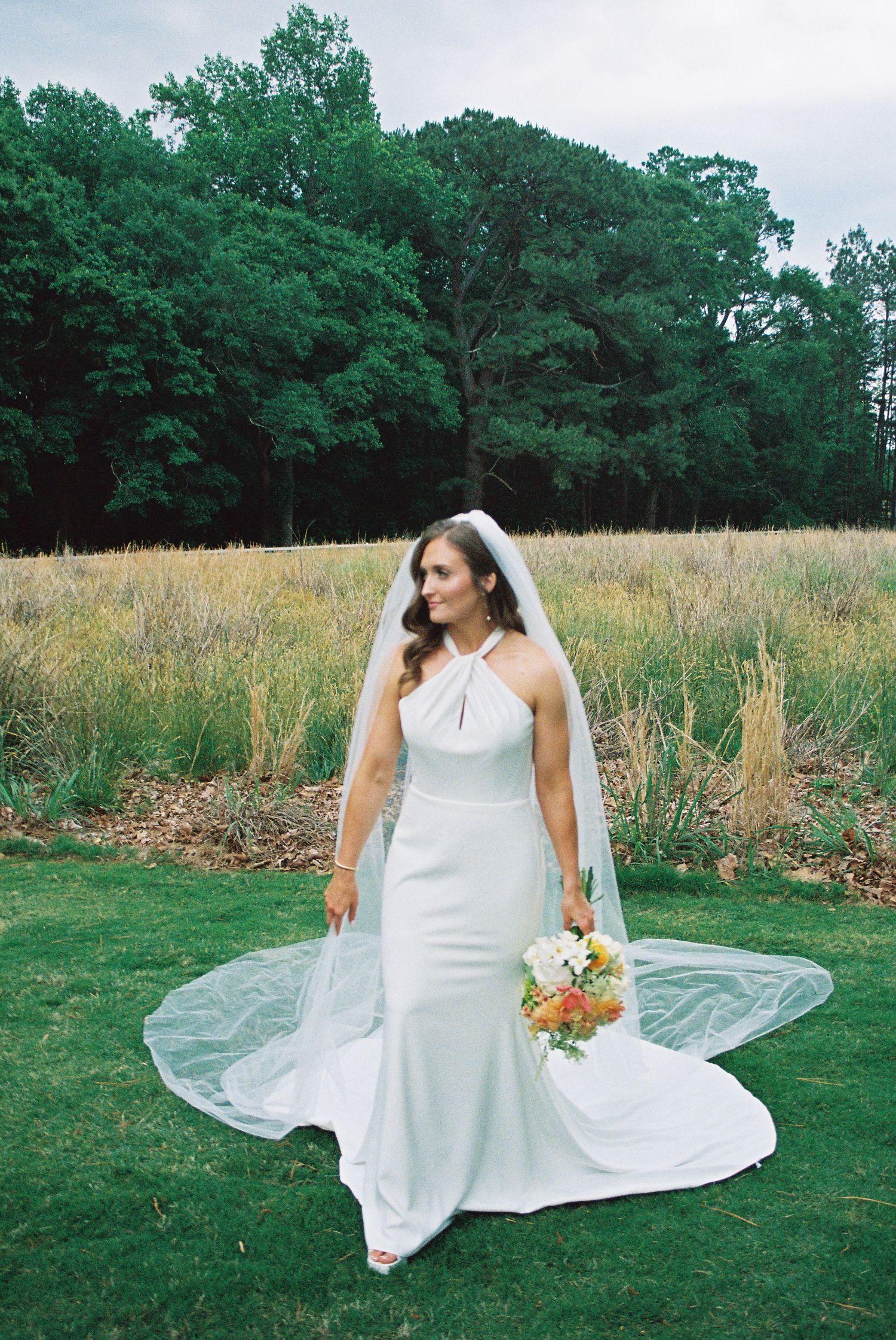 Bride in white halter wedding gown with veil holding colorful bouquet standing on green lawn with field and trees behind.