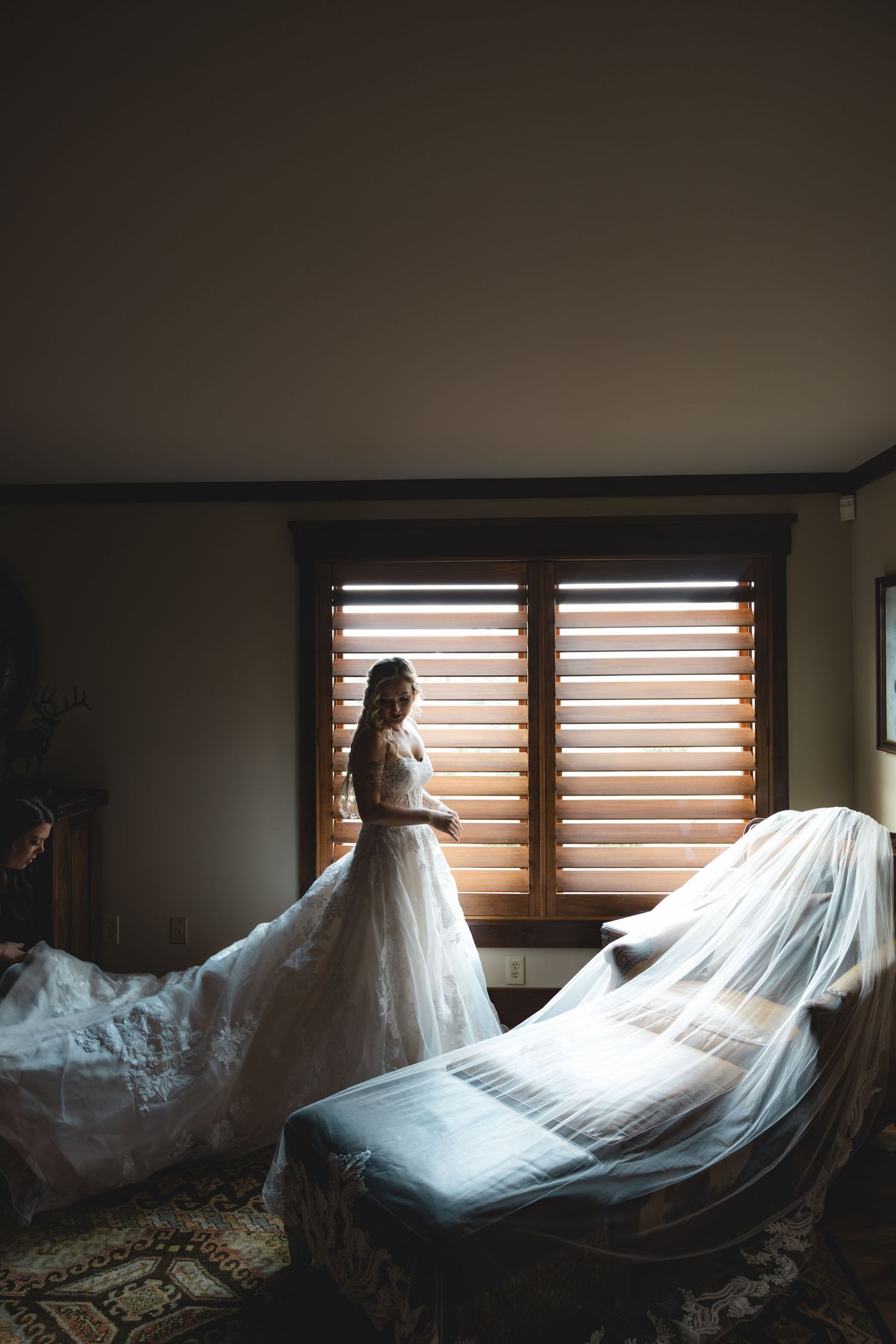 Bride in white lace gown stands by window as assistant arranges flowing veil and train in dimly lit room.