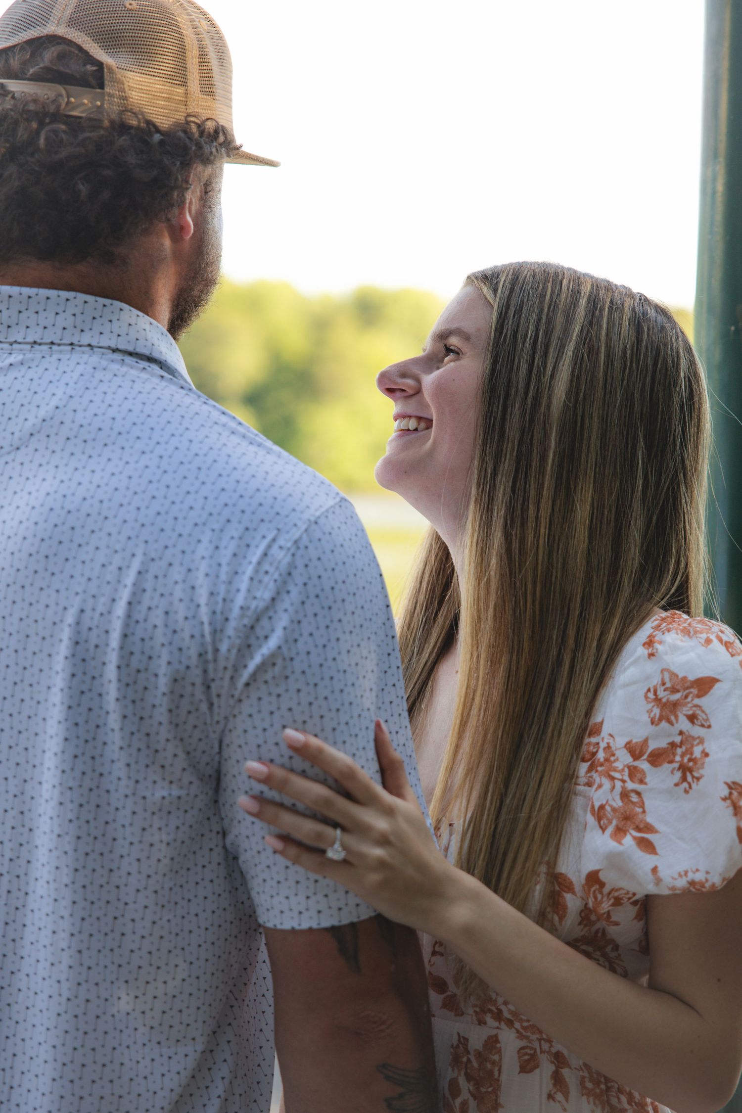 Smiling woman in floral dress gazes at man in baseball cap, showing engagement ring outdoors.