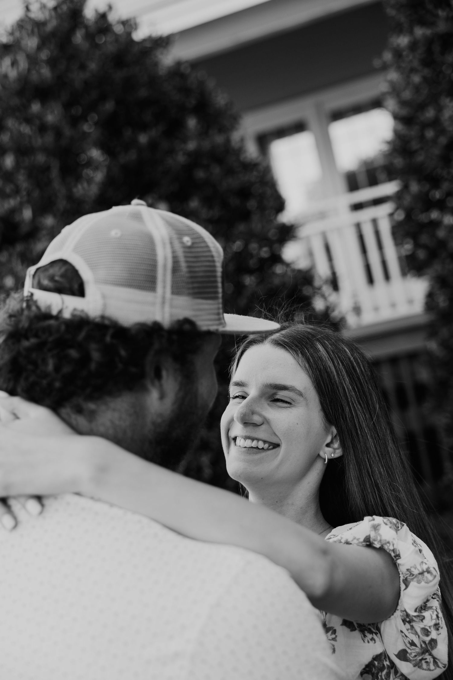 Black and white photo of a smiling woman hugging a man in a trucker hat outdoors near a house.