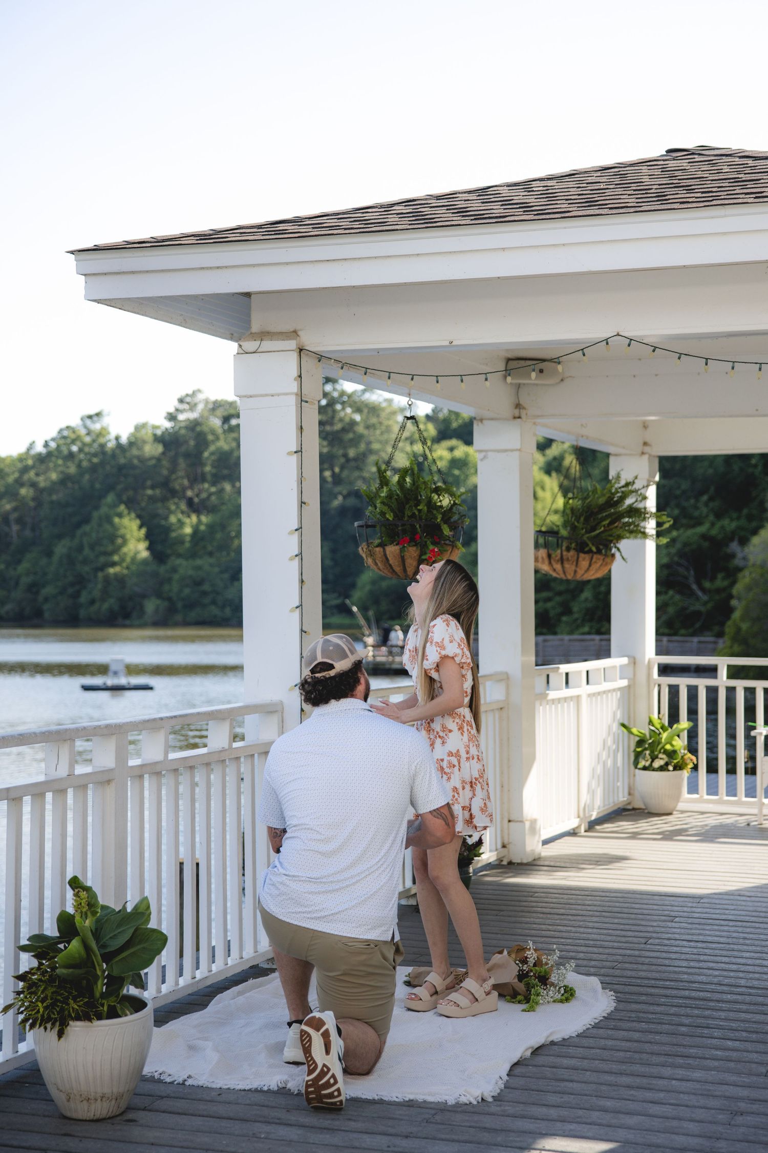 Man kneeling on gazebo deck proposing to woman in floral dress by lakeside, romantic outdoor marriage proposal.