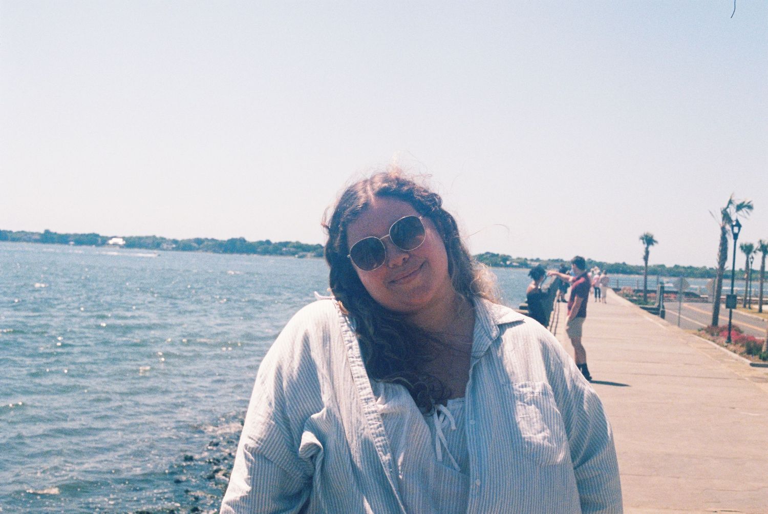 Smiling woman in sunglasses and light blue jacket stands on a sunny waterfront promenade by the sea.