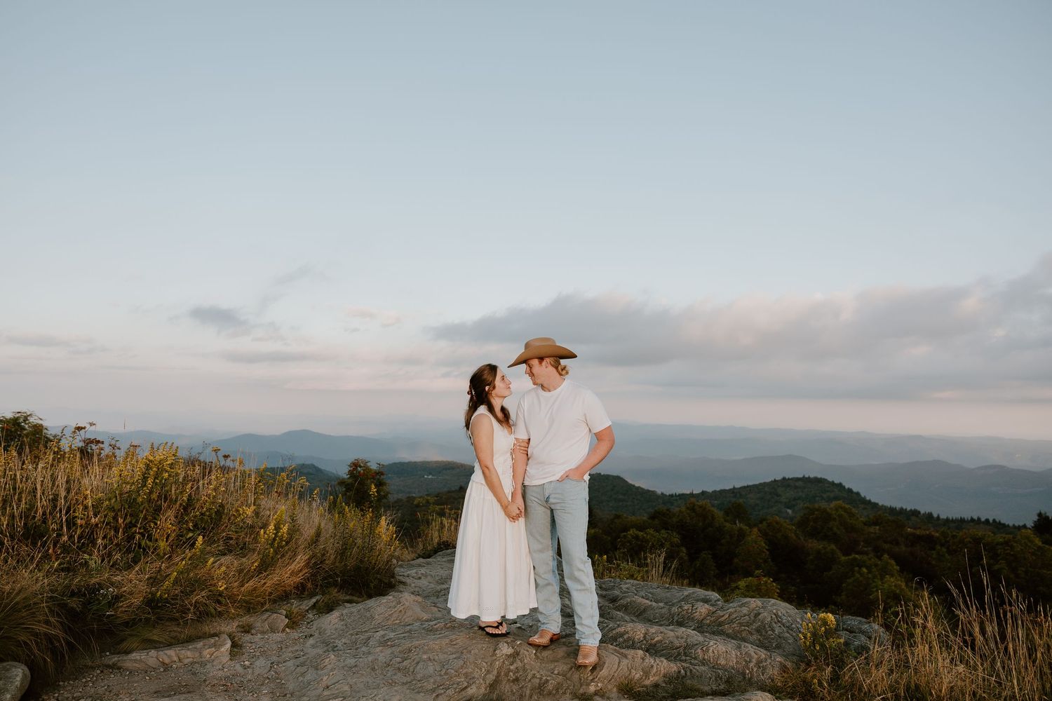Couple embracing on a mountaintop at sunset with scenic mountain views in the background.