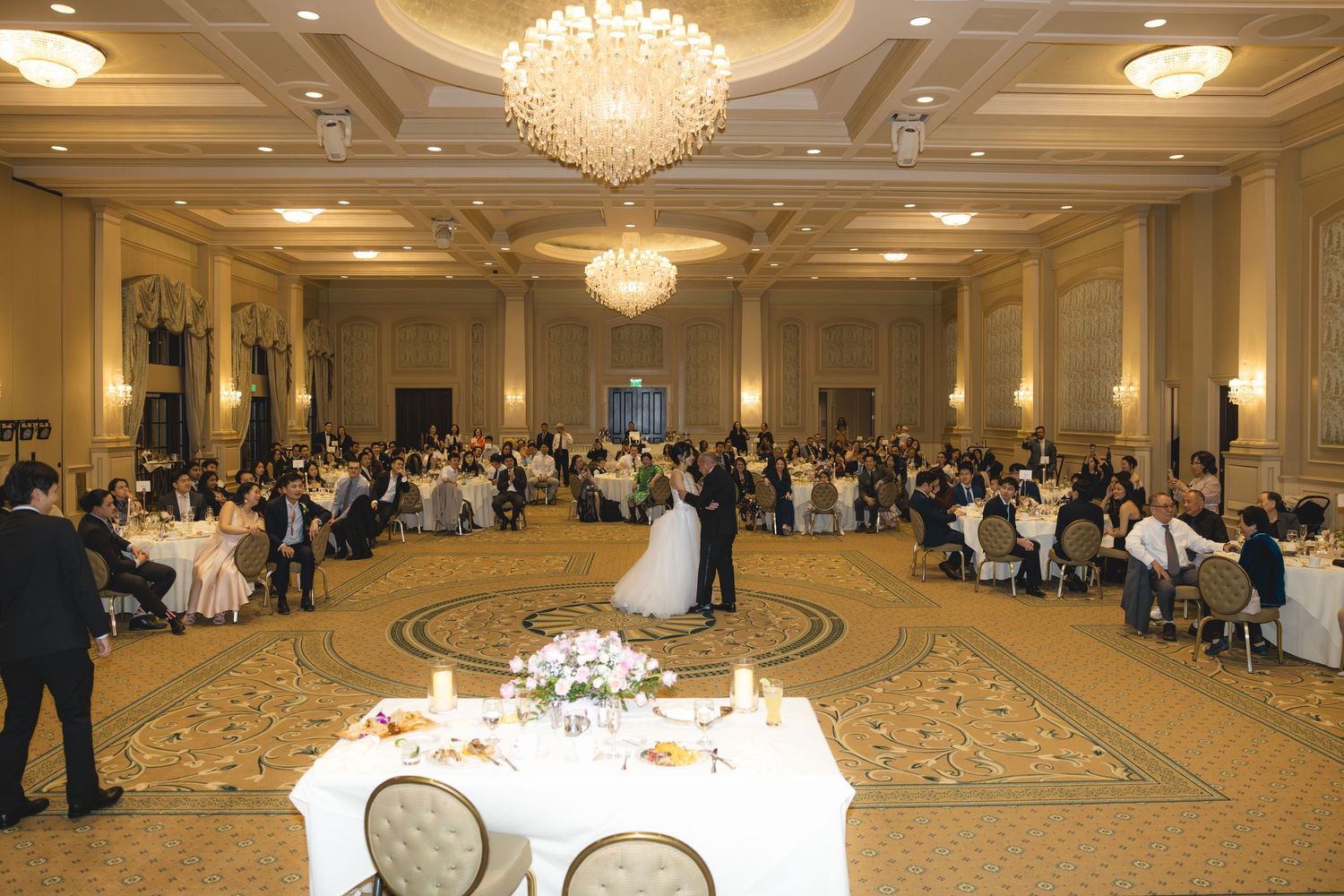 Bride and groom share first dance in elegant chandelier-lit wedding reception ballroom.
