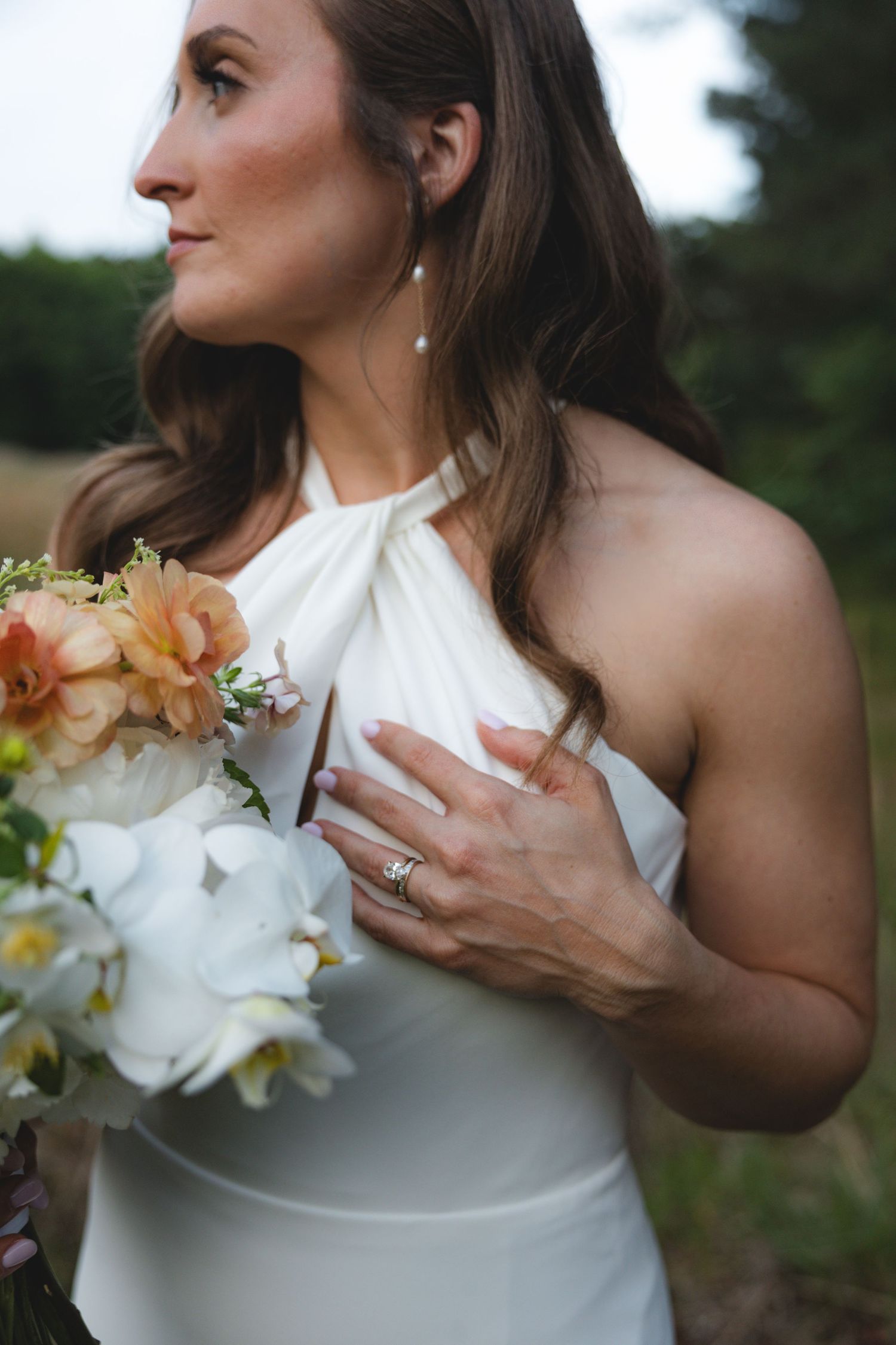 Close-up of bride in white halter gown holding peach and white bouquet with pearl drop earrings outdoors.