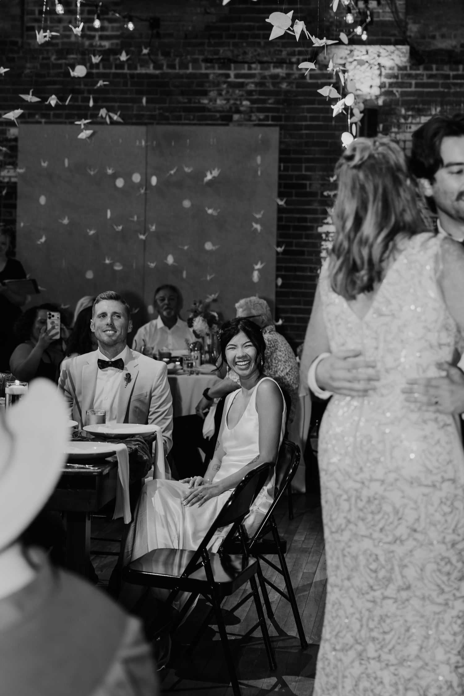 Black and white wedding reception photo of smiling bride seated with groom in bow tie, guests celebrating in elegant venue.