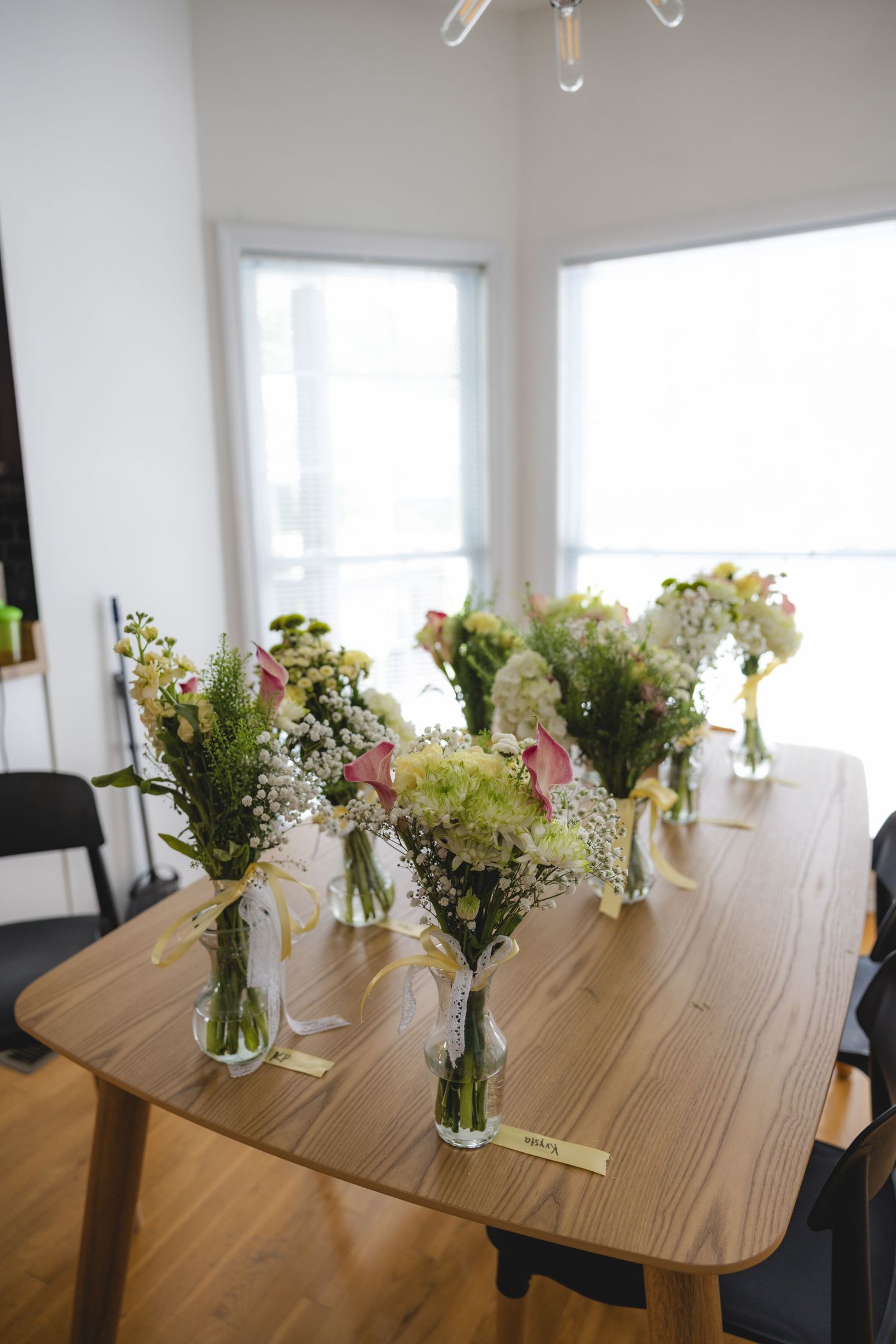Multiple floral bouquets in glass vases with yellow ribbons arranged on a wooden dining table near bright windows.