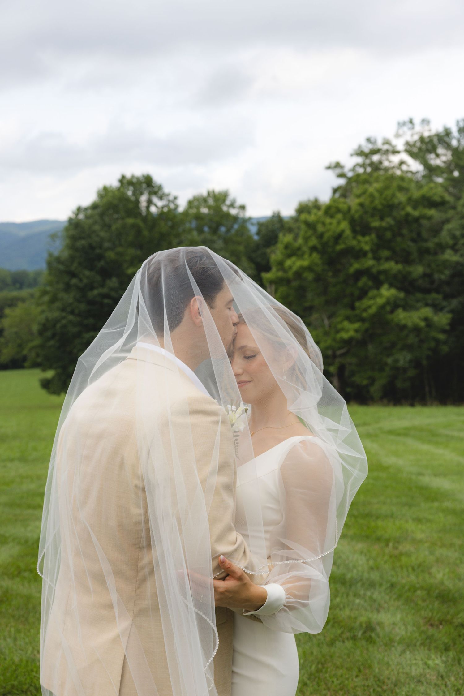 Bride and groom share intimate moment under wedding veil in lush green outdoor setting.