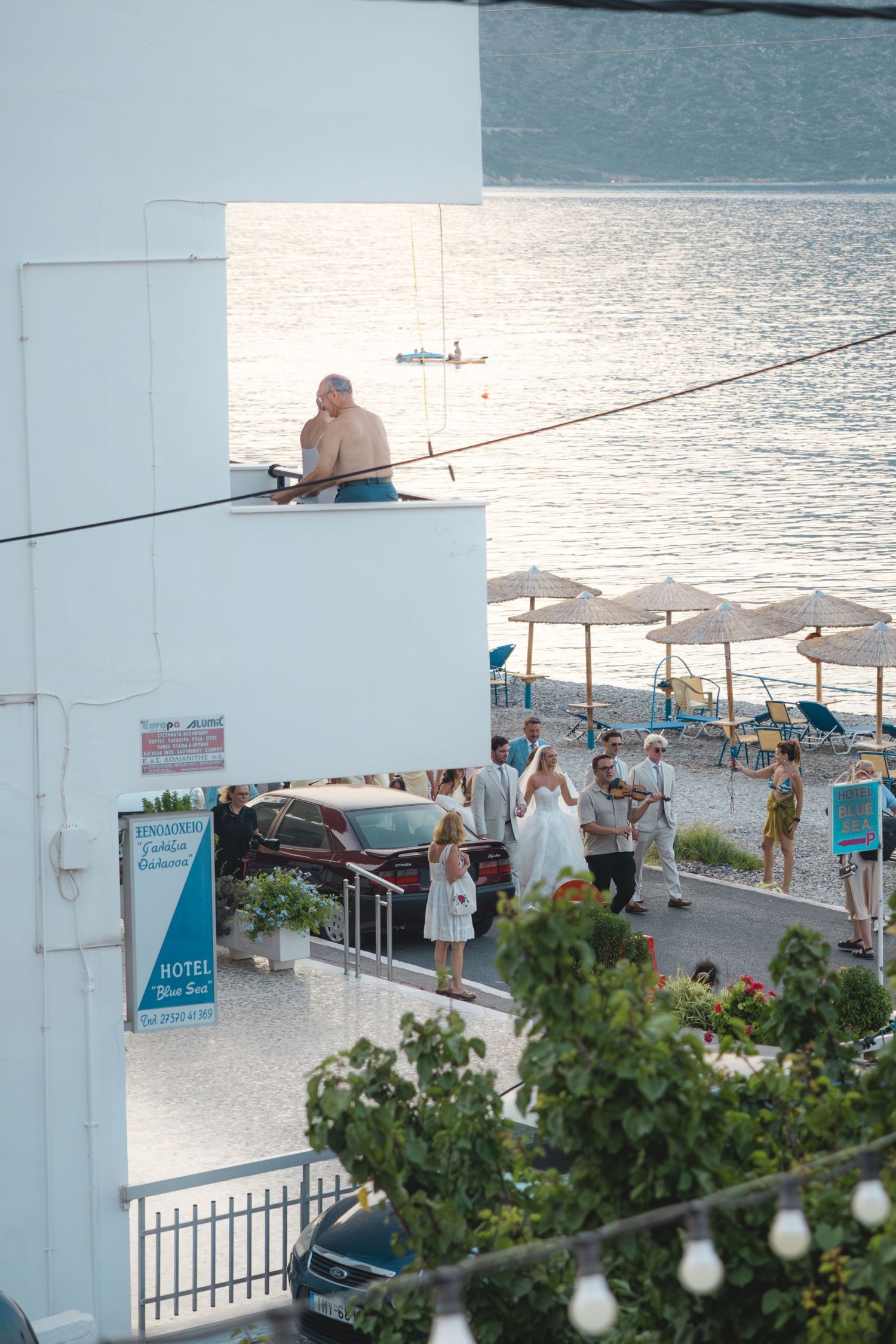 Coastal hotel scene with wedding party gathering near beachfront, umbrellas and calm sea visible in background.
