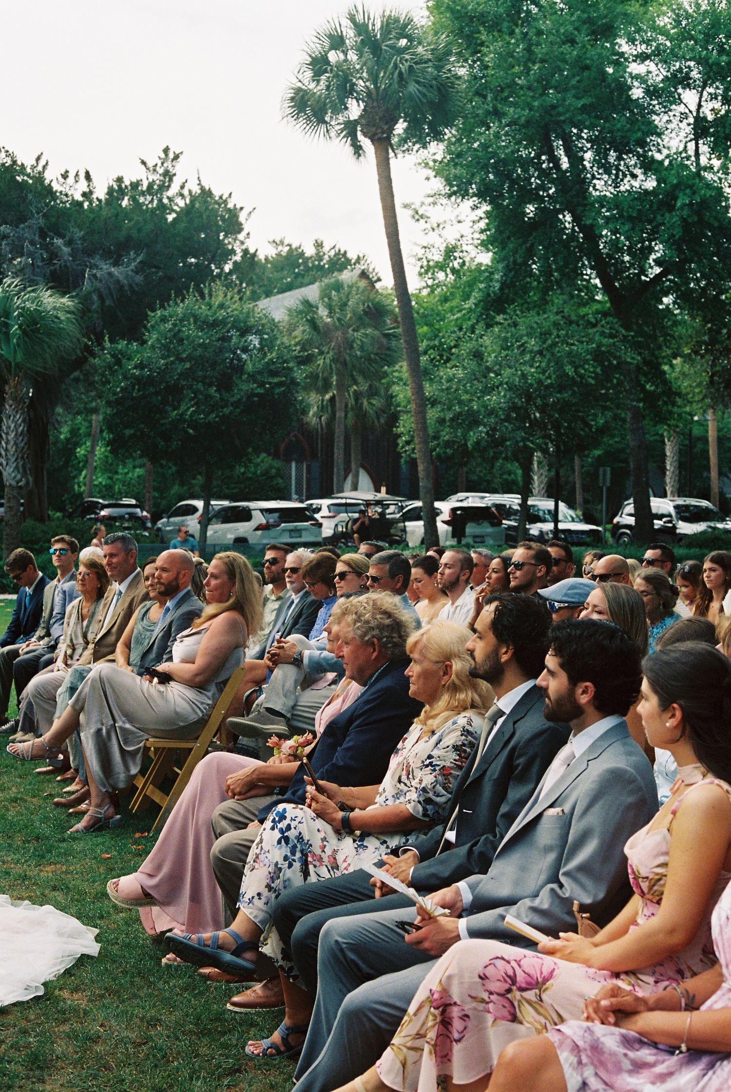 Outdoor wedding ceremony guests seated on chairs surrounded by palm trees and lush greenery.