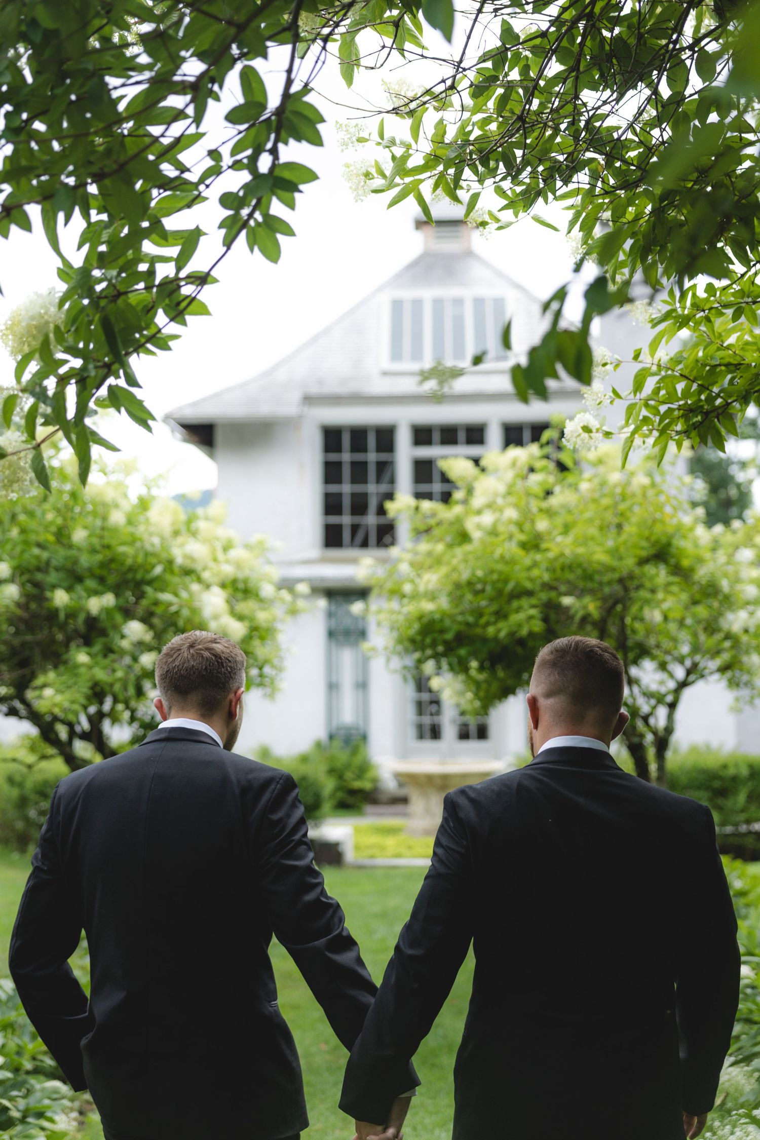 Two grooms in black suits hold hands in a lush garden, facing a white house on their wedding day.
