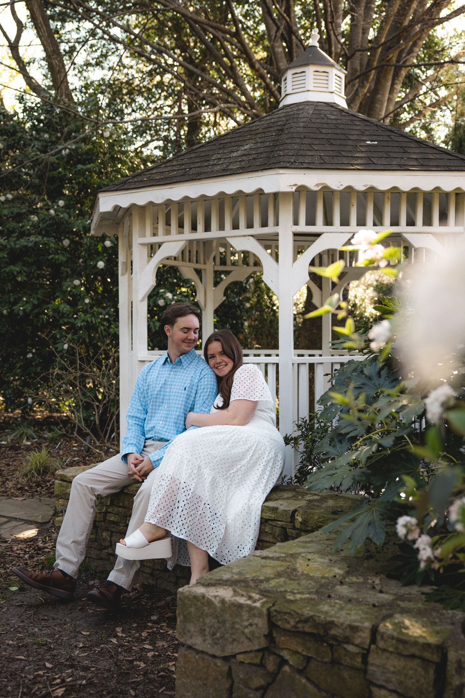 Couple sitting together on a stone wall in front of a white gazebo in a lush garden setting.