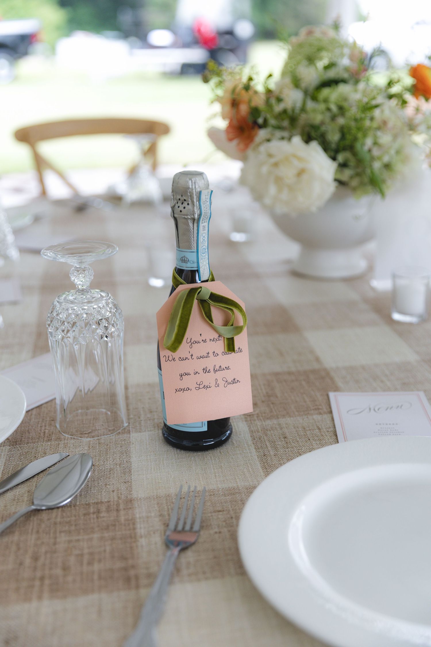 Mini champagne bottle wedding favor with handwritten pink tag and green ribbon on plaid tablecloth.