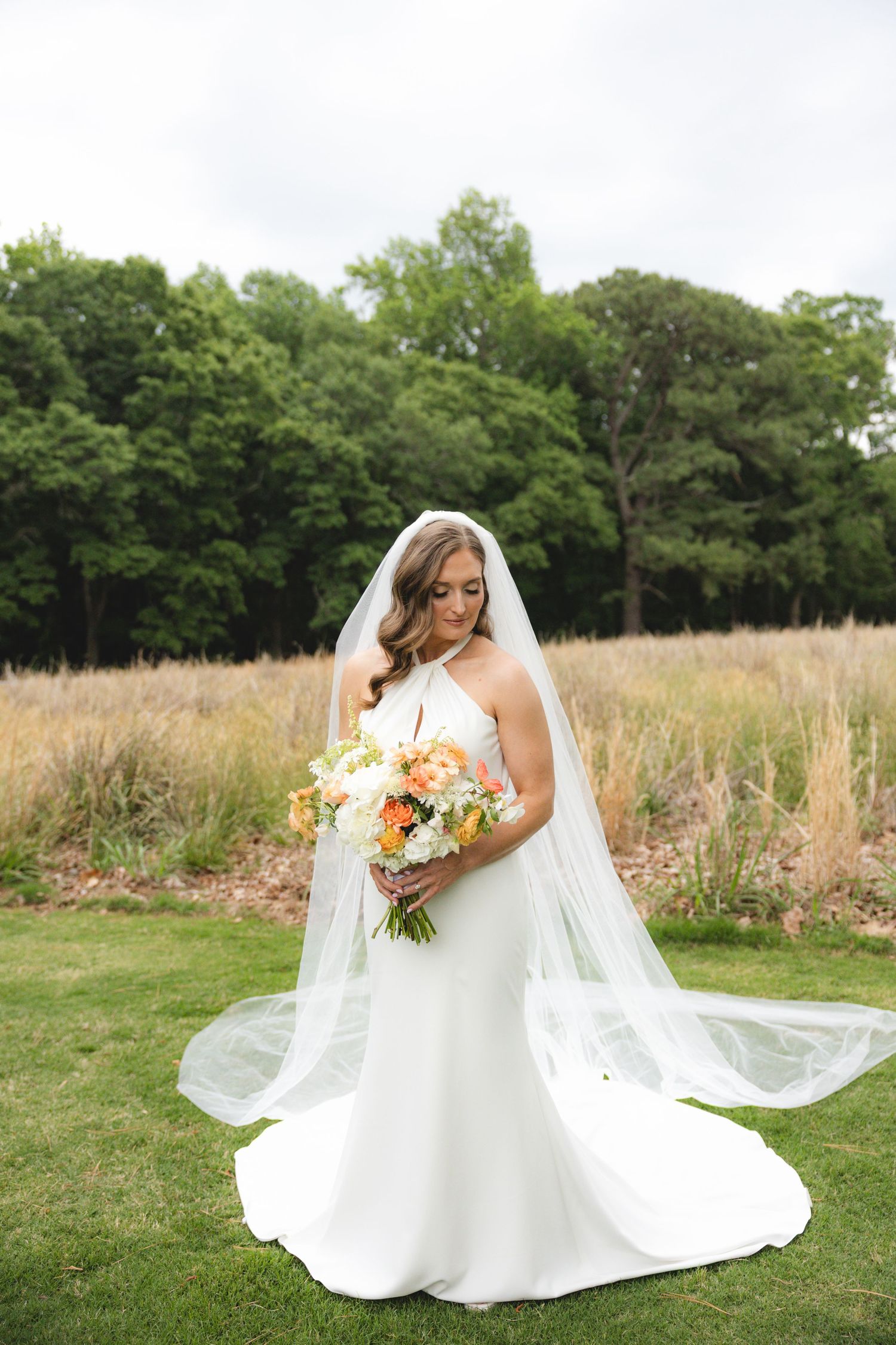 Bride in white halter gown and veil looks down at peach and white bouquet standing on green lawn outdoors.