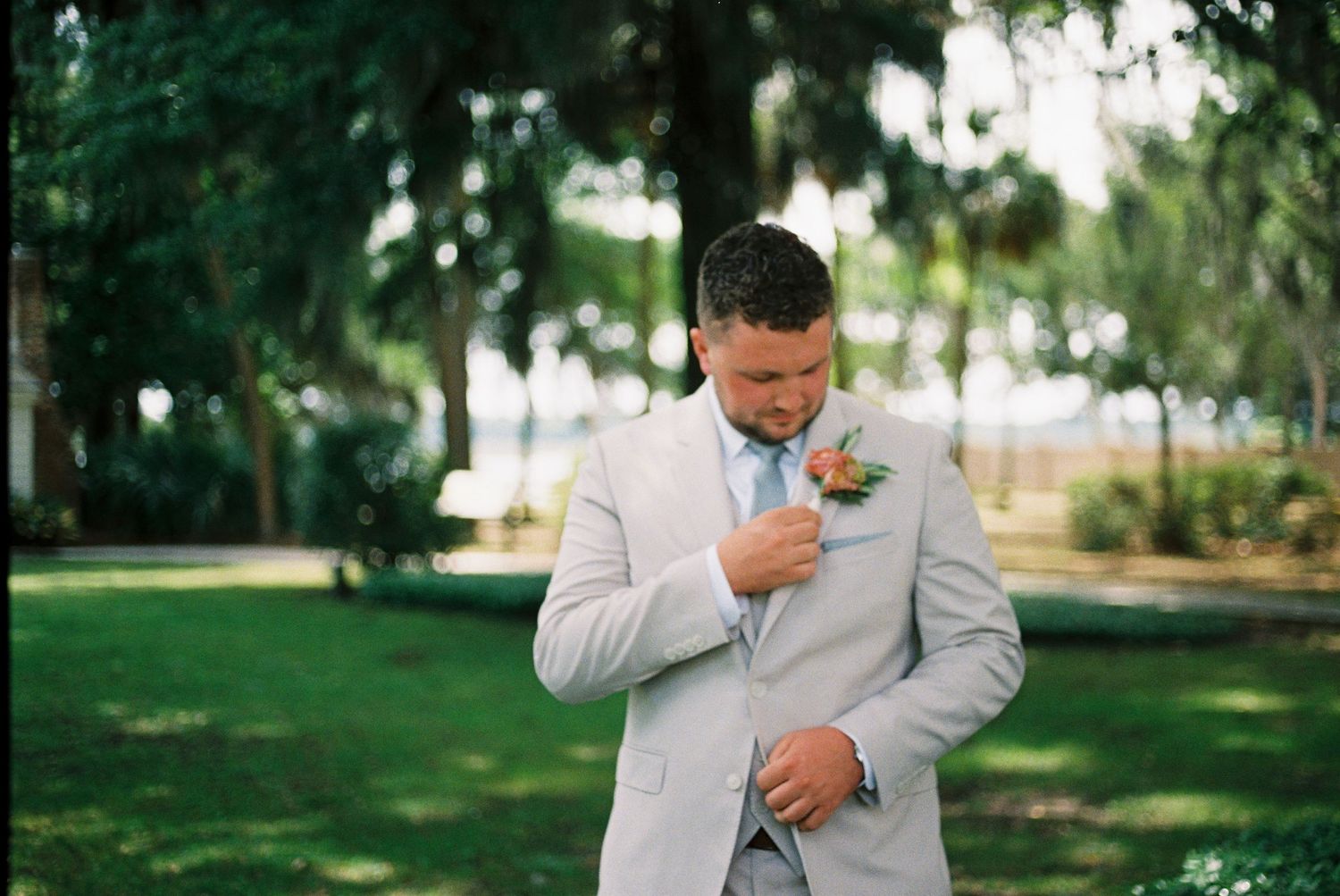 Handsome groom in light gray suit adjusting boutonniere in lush green outdoor garden setting.