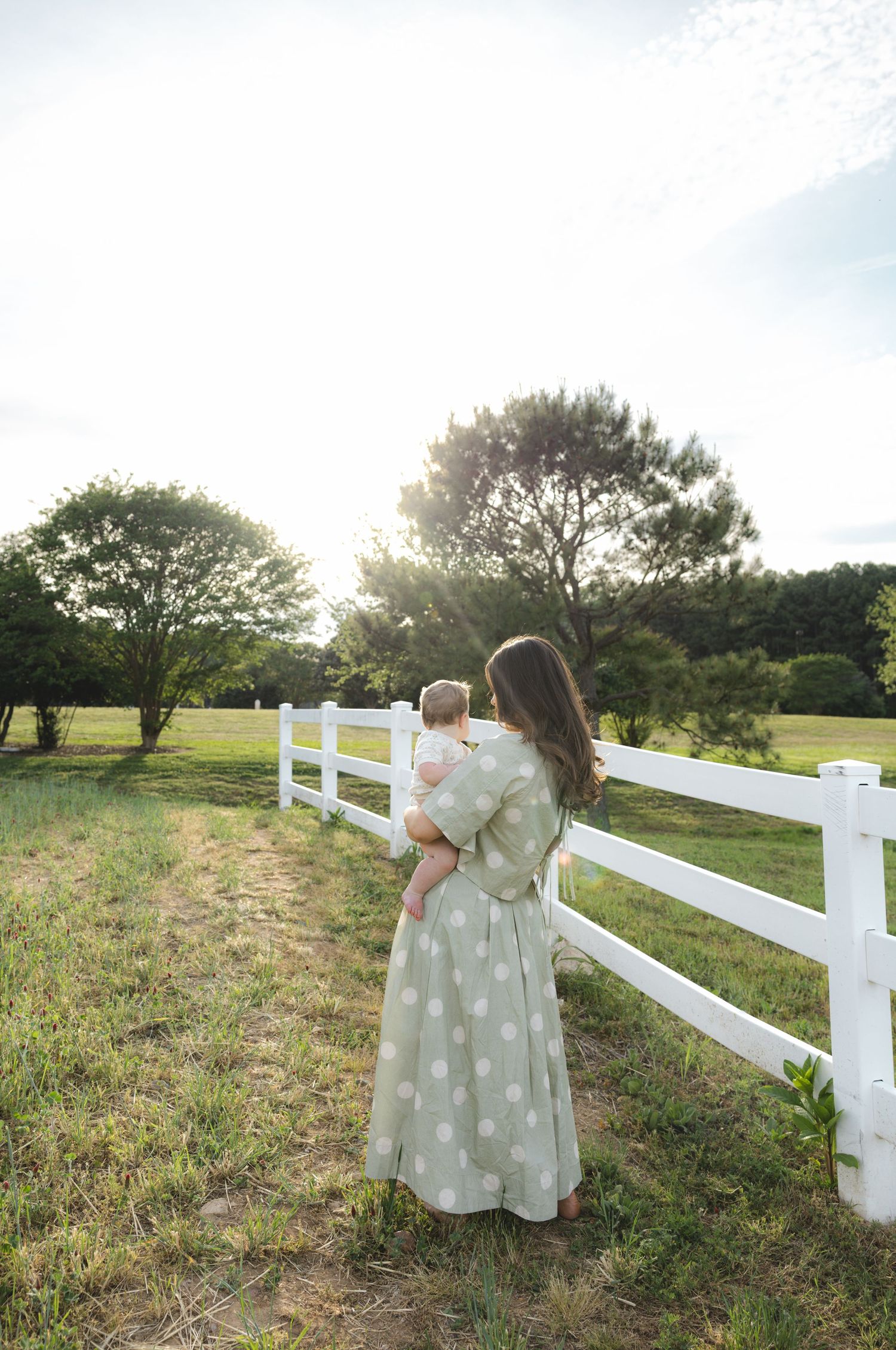 A mother in a polka dot dress holds her baby by a white fence at golden hour on a lush green farm.