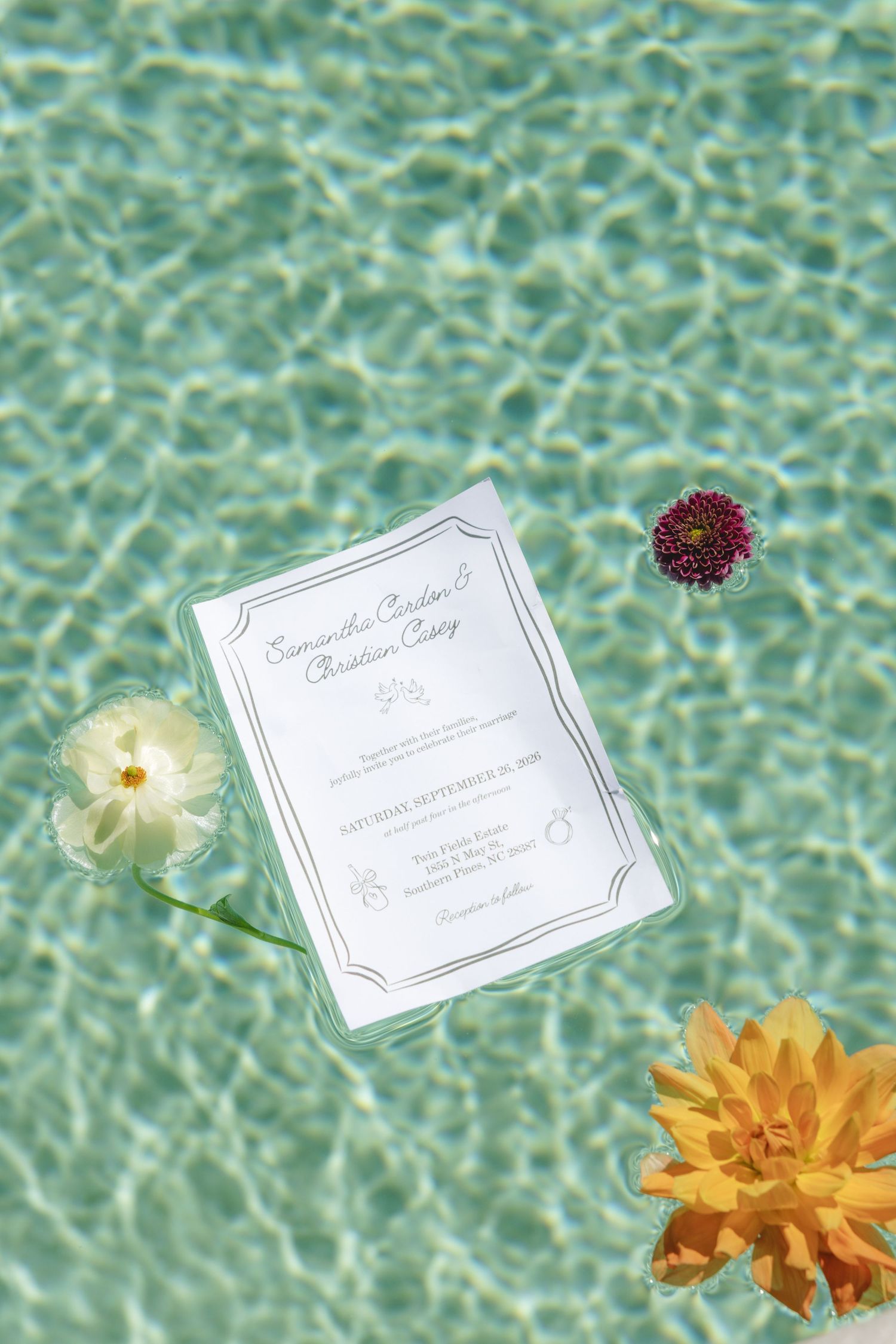Wedding invitation floating in turquoise pool water surrounded by white, yellow, and burgundy flowers.