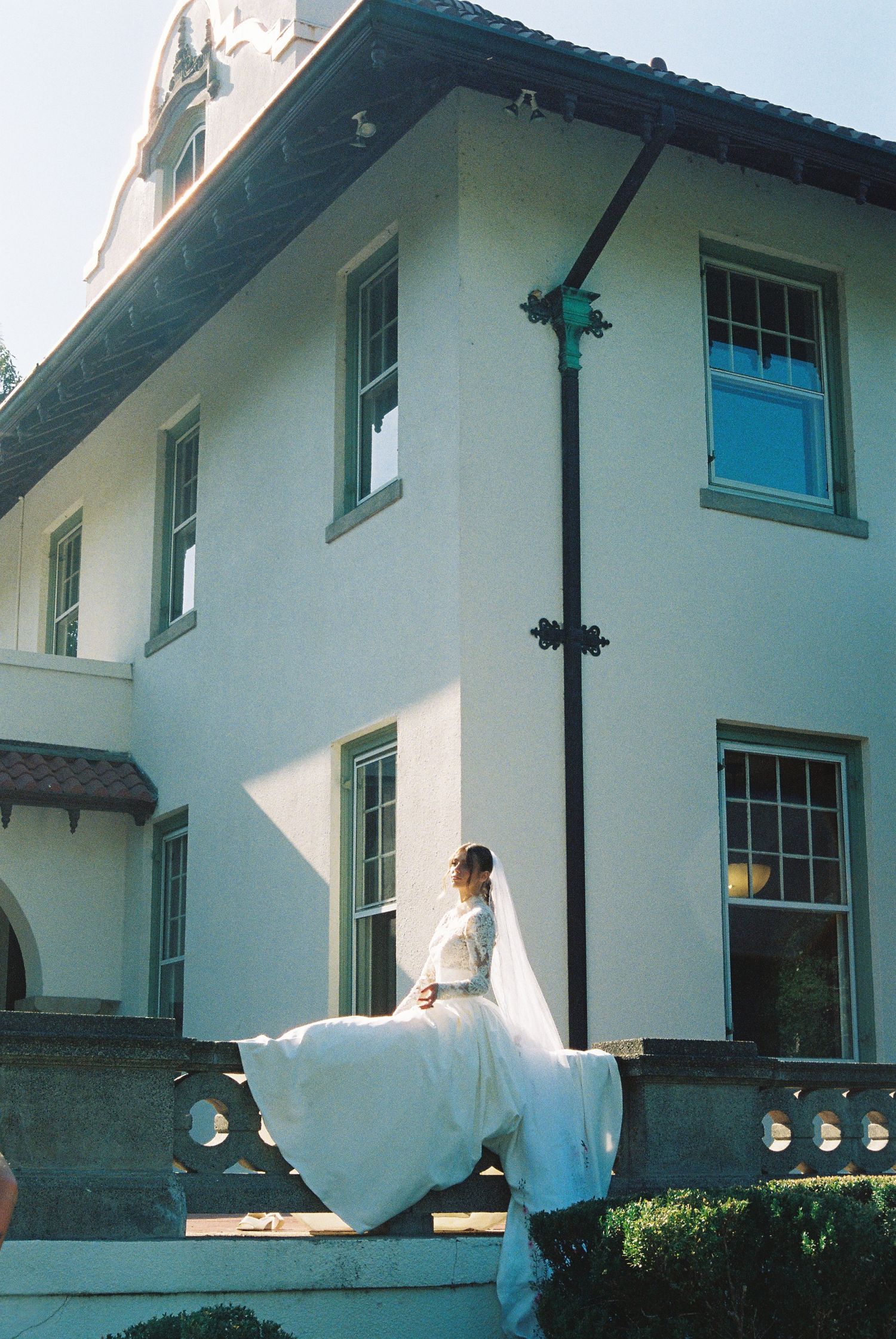 Bride in flowing white gown and veil poses on stone ledge outside elegant white mansion on sunny day.