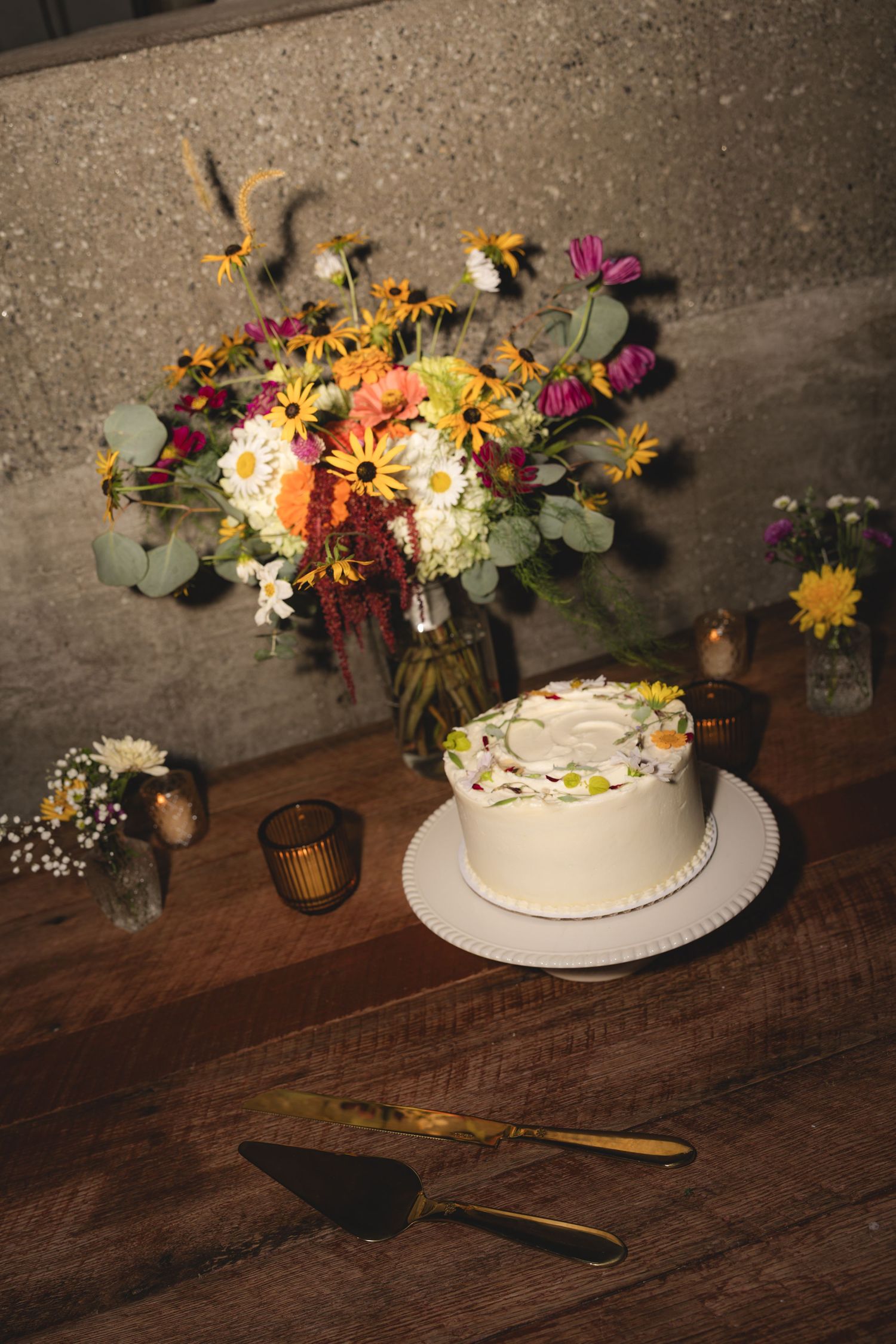 White frosted wedding cake with floral decorations beside a colorful wildflower bouquet on a rustic wooden table.