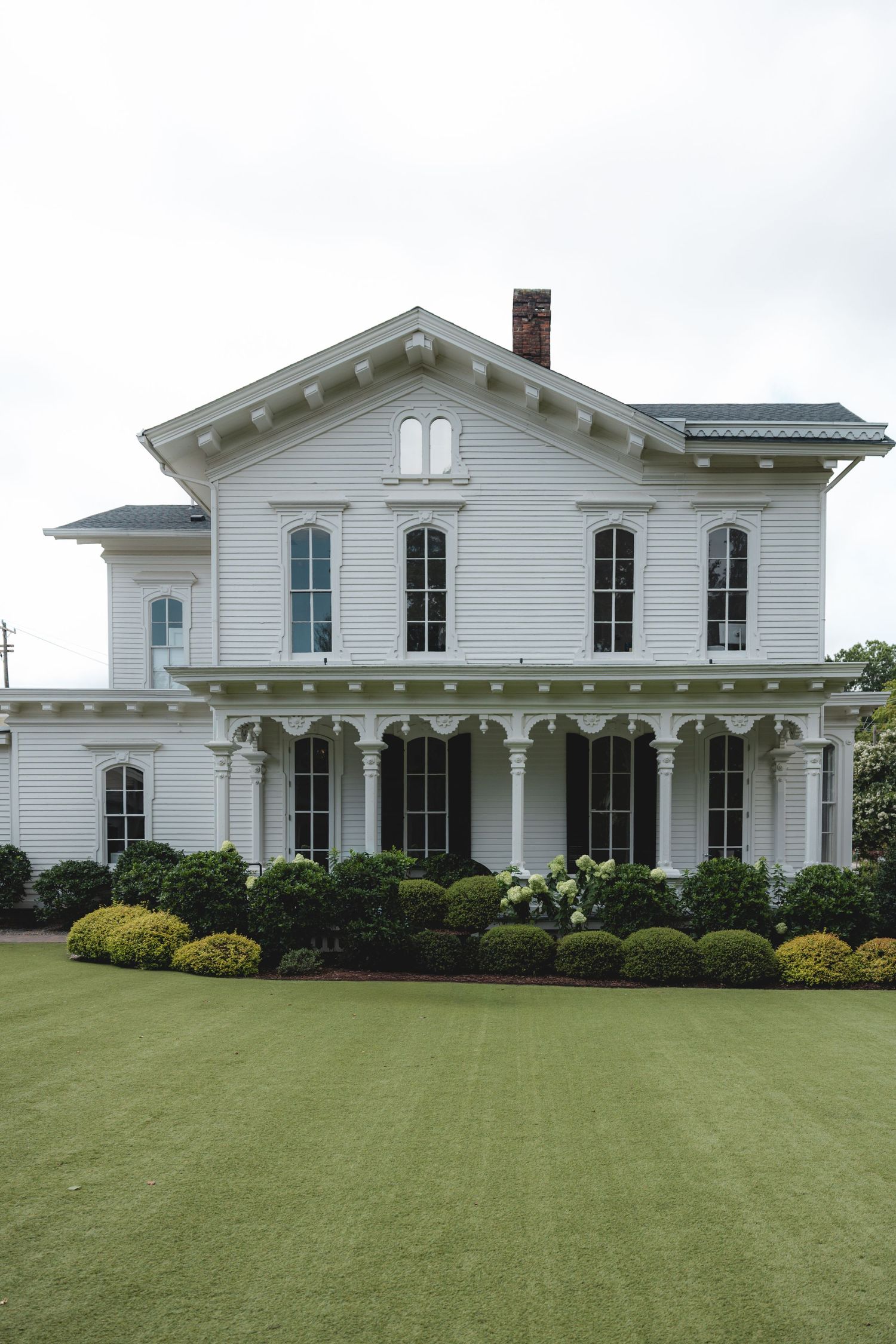 White two-story Victorian home with covered porch, decorative trim, manicured shrubs, and lush green lawn on overcast day.