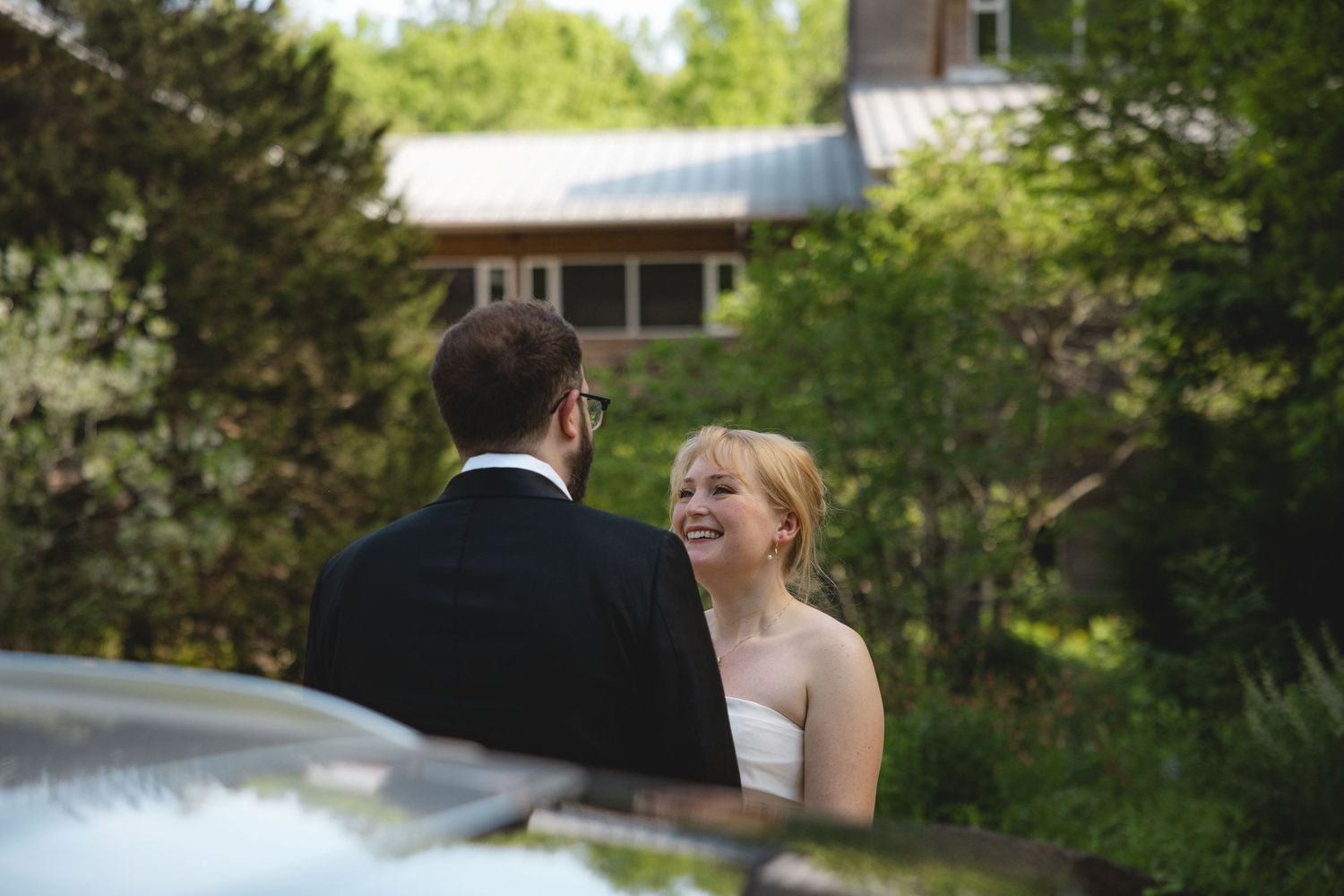 Bride in white dress laughing joyfully with groom in black suit outdoors near rustic building surrounded by lush greenery.