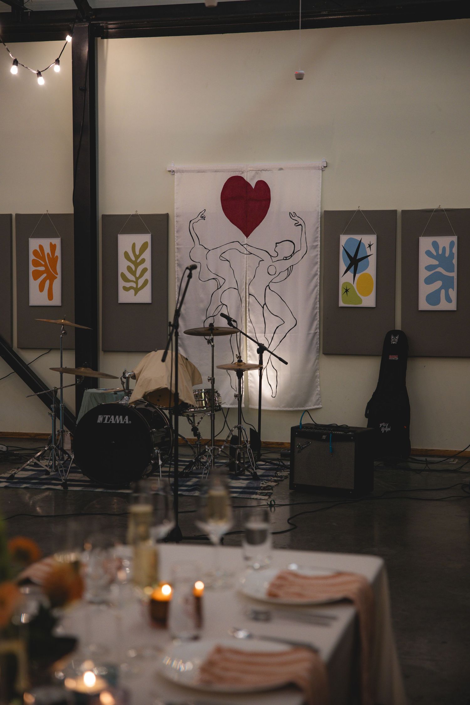 Elegant wedding reception venue with drum kit, heart artwork banner, colorful paintings, and candlelit dining tables.