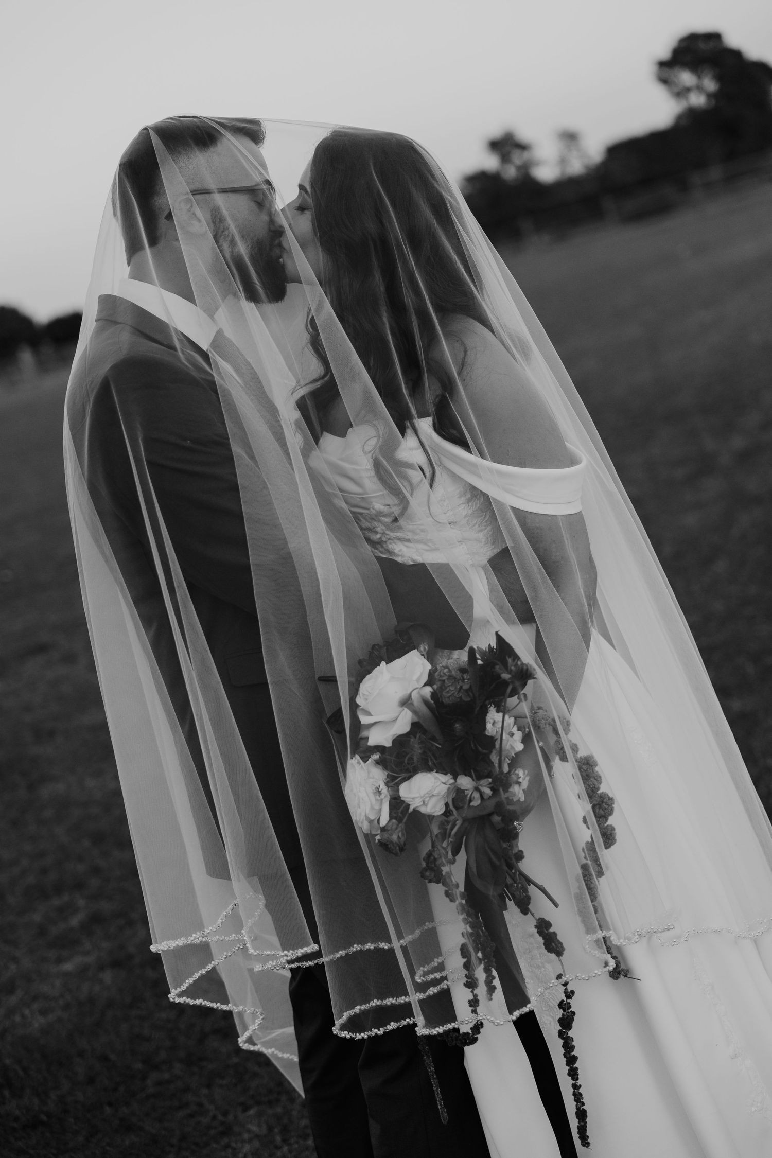 Black and white photo of bride and groom kissing under a wedding veil outdoors, bride holding a bouquet.