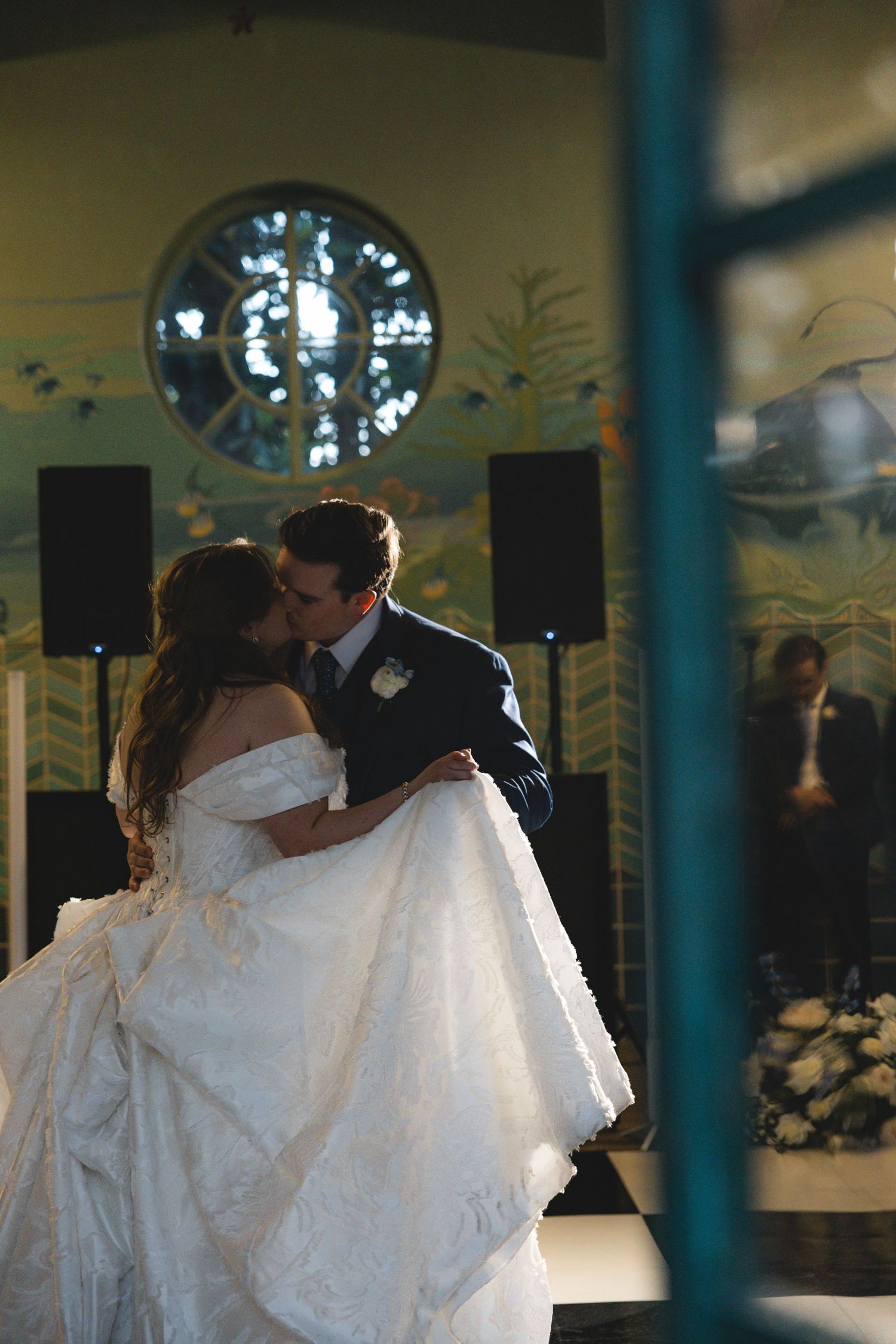 Bride and groom share a romantic first dance kiss in an elegant venue with stained glass window backdrop.