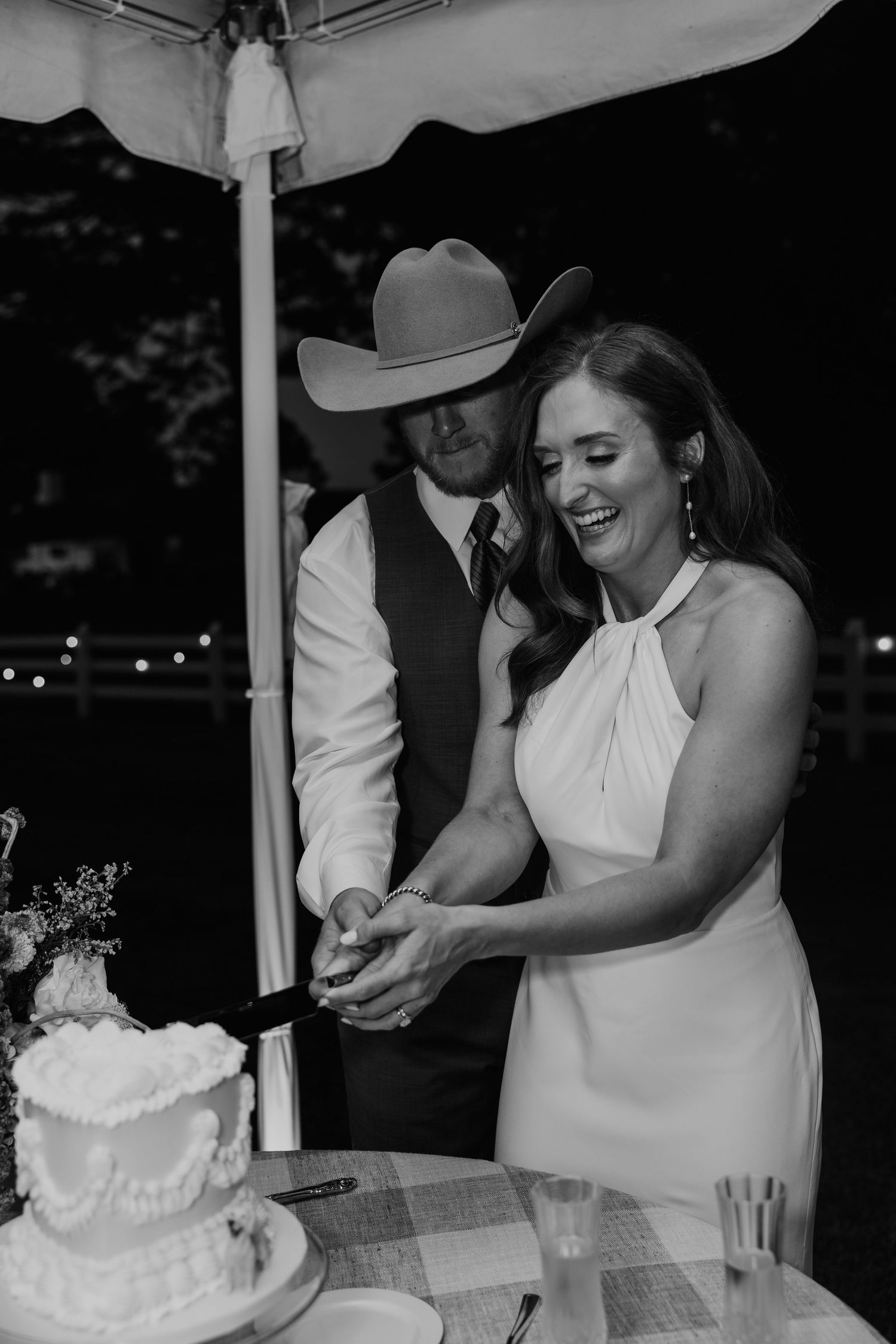 Black and white photo of smiling bride and groom in cowboy hat cutting wedding cake together.