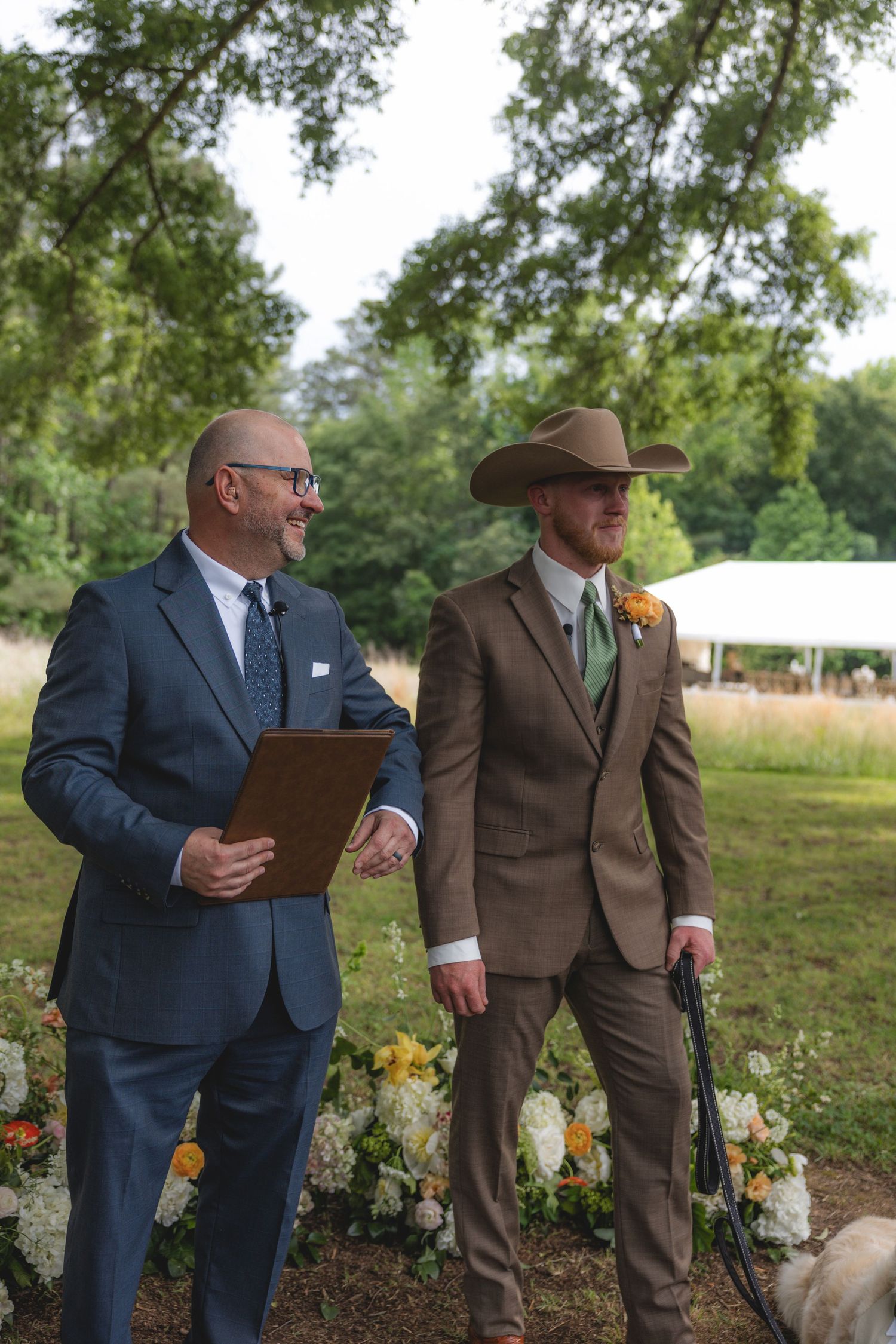 Groom in brown suit and cowboy hat stands with officiant in navy suit at outdoor wedding ceremony.