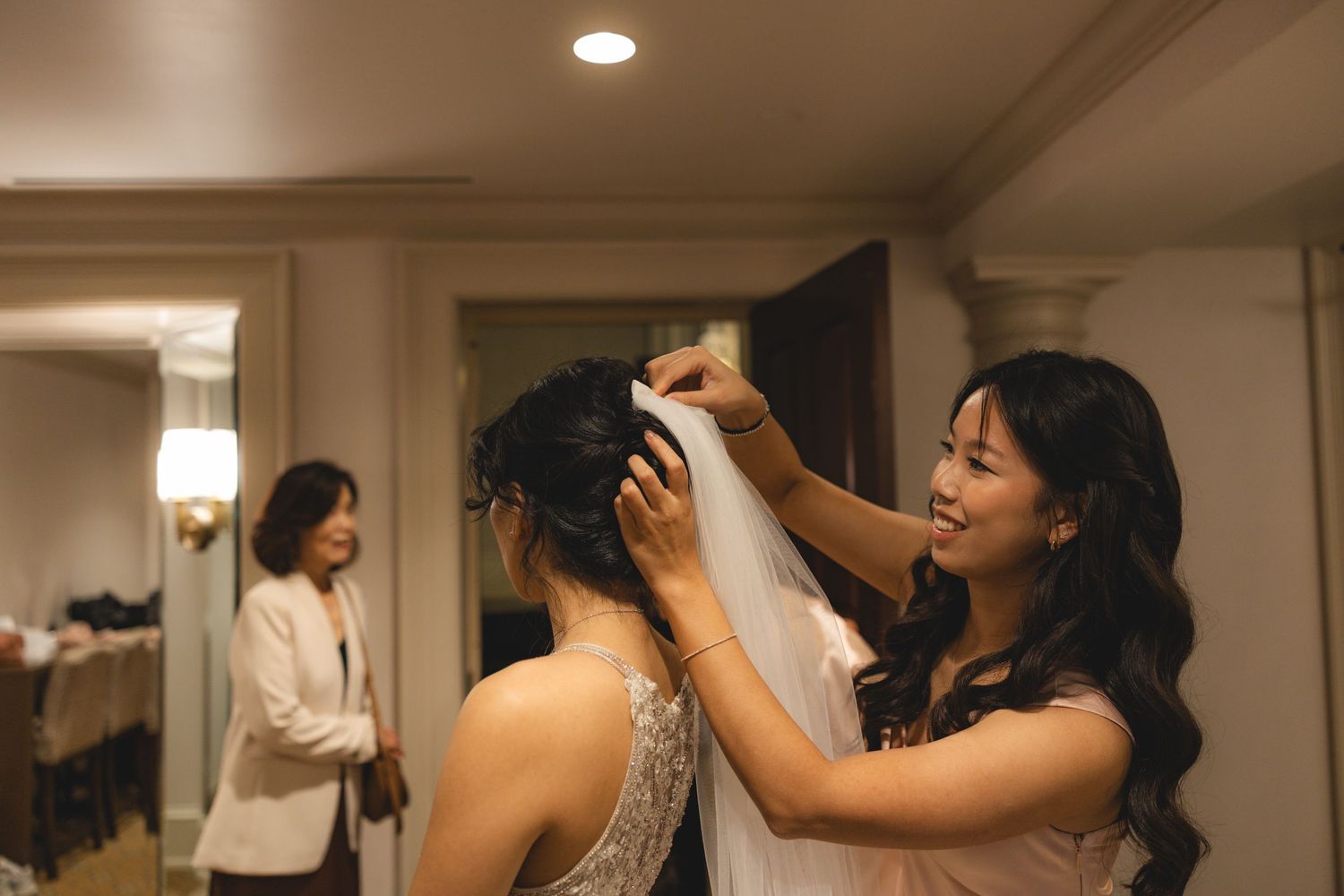 Smiling bridesmaid carefully adjusts bride's veil while another guest watches in the background during wedding preparations.