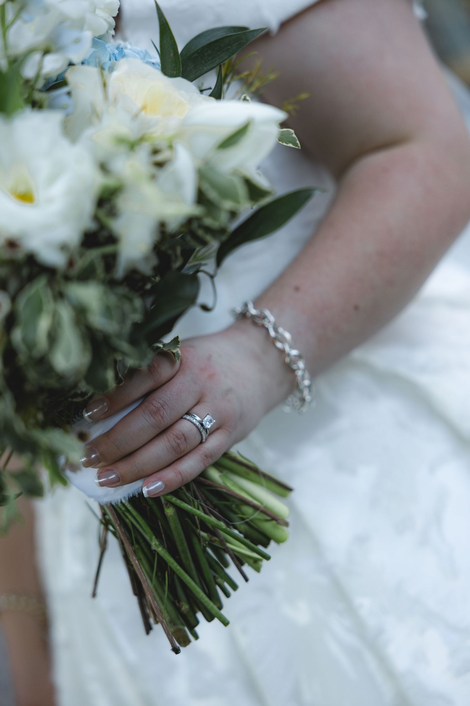 Bride holding white floral wedding bouquet with diamond ring and silver bracelet on wrist in white gown.