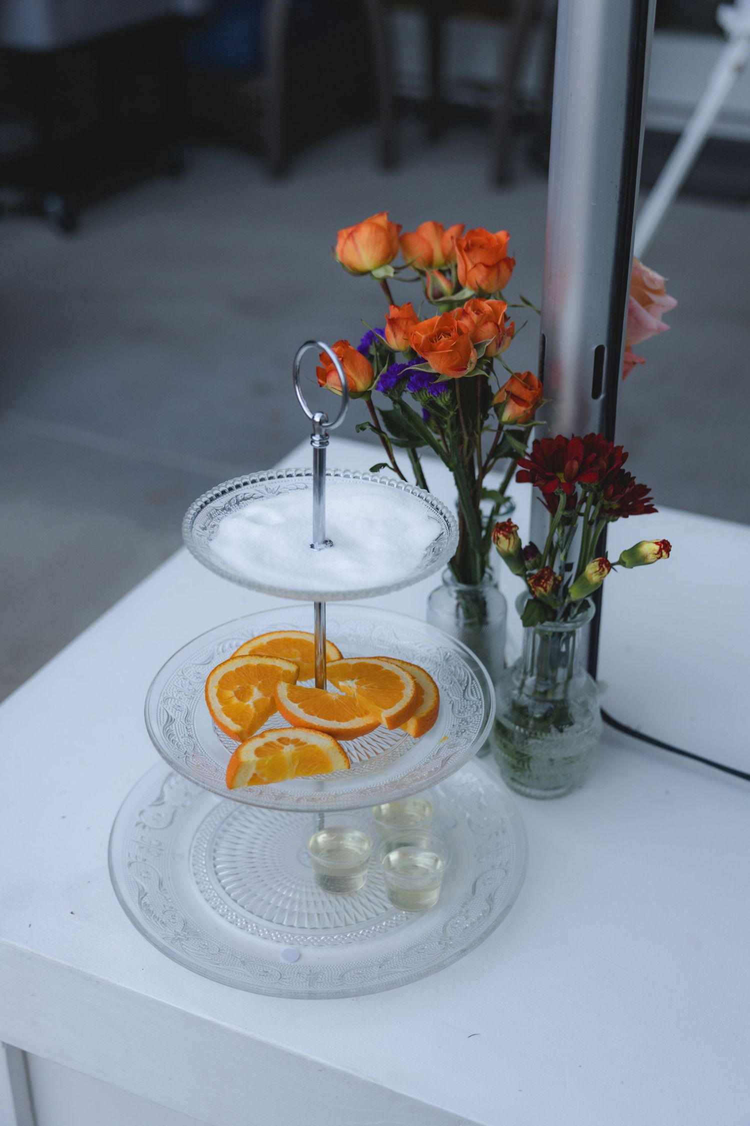 Three-tier glass serving stand with orange slices and small glasses, beside vases of orange and red flowers on white table.