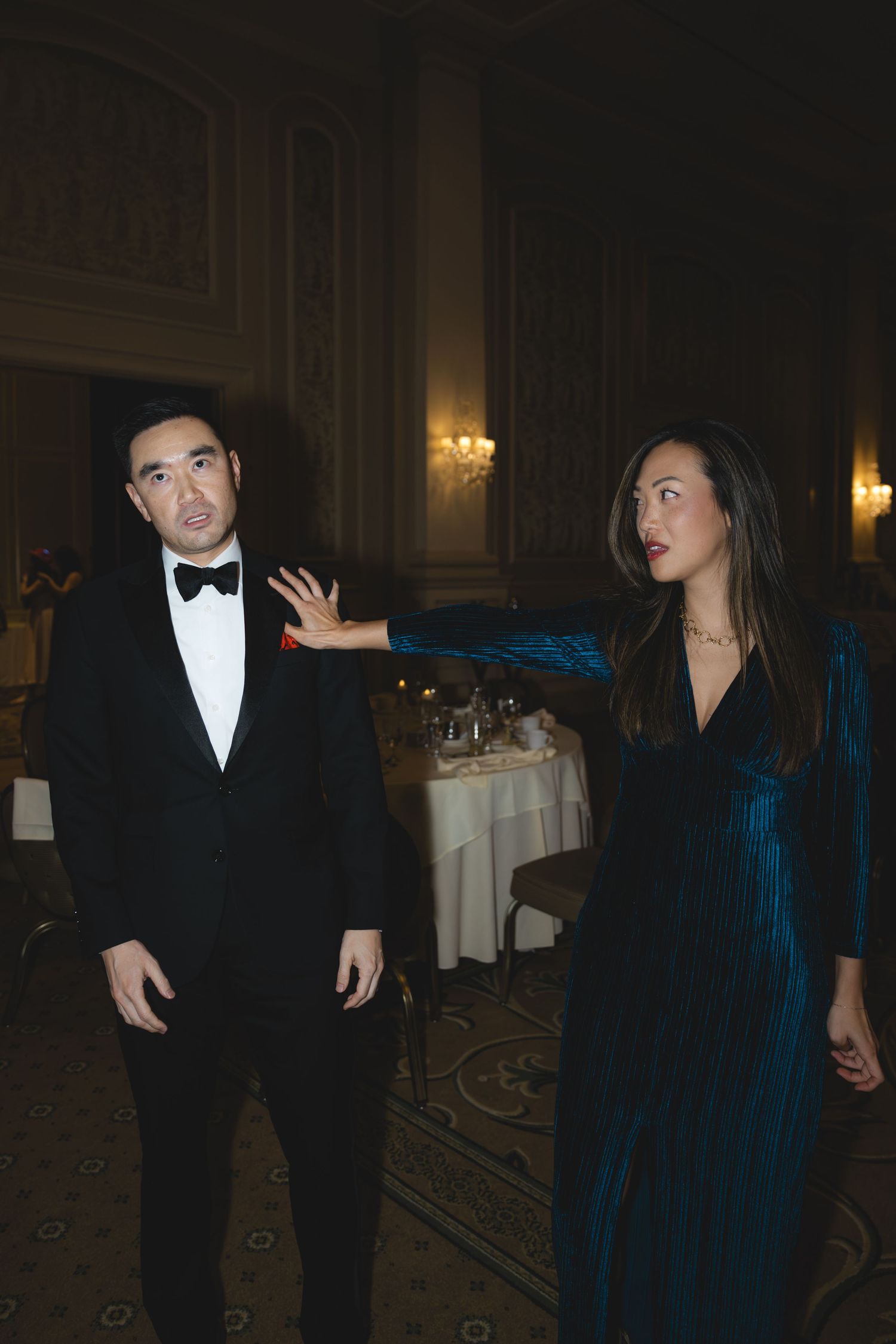 Elegant couple in formal attire at a wedding reception, woman in teal velvet gown gesturing to man in tuxedo.