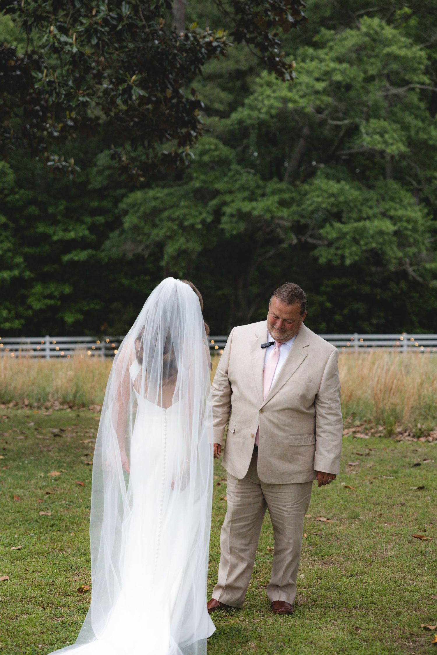 Father of the bride in tan suit walking alongside bride in white gown and cathedral veil outdoors.