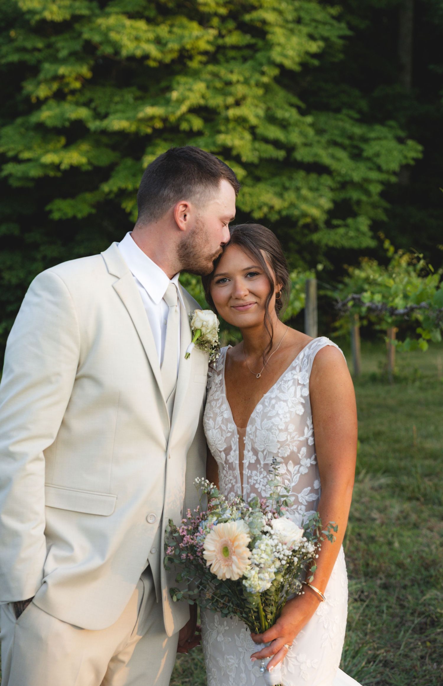 Groom in beige suit kissing bride's forehead as she smiles holding a white floral bouquet outdoors.