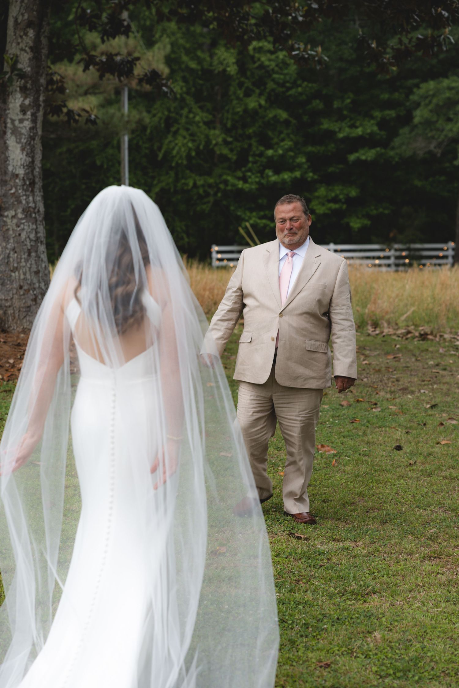 Bride in white gown with long veil stands in a field as a man in a tan suit approaches during first look.