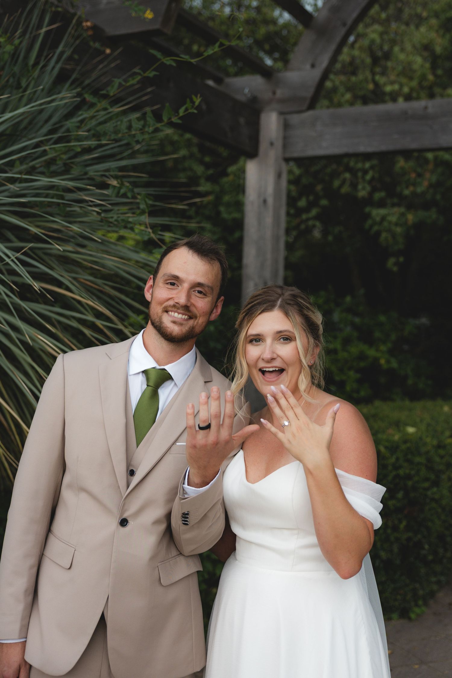 Smiling newlyweds in garden showing off wedding rings, groom in tan suit green tie, bride in white off-shoulder gown.