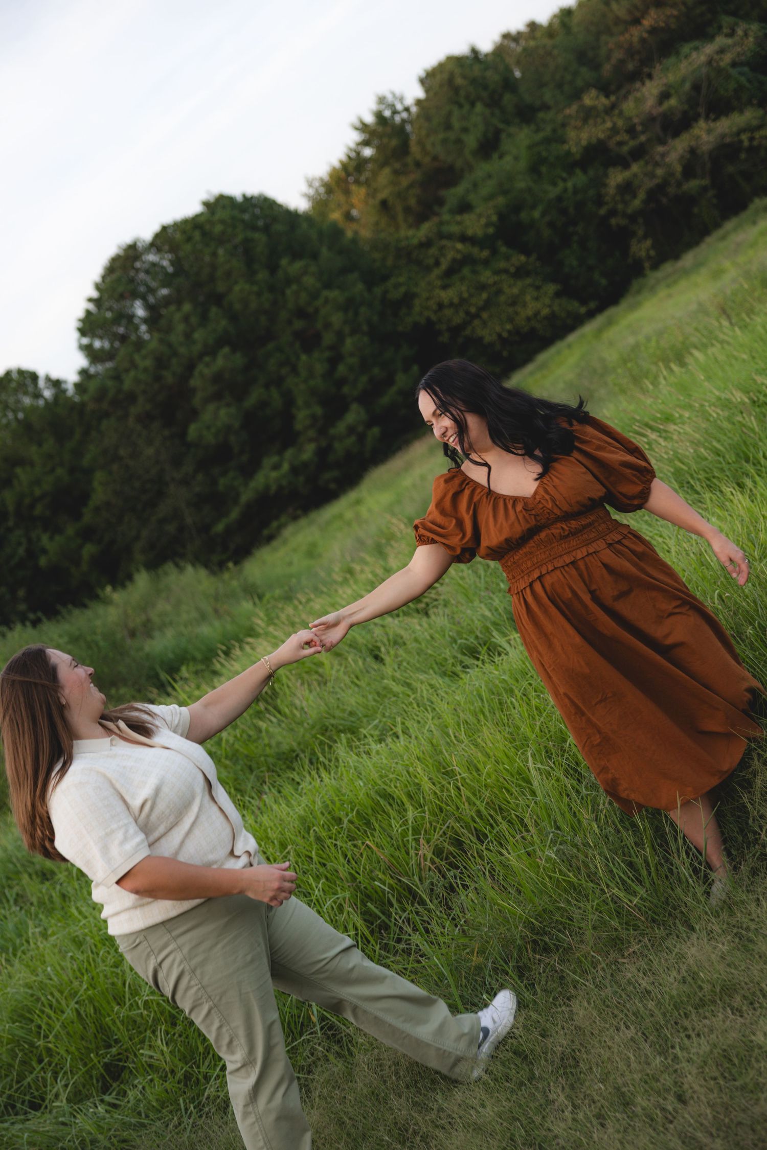 Two women holding hands on a green grassy hillside, one in a brown dress and one in white shirt and khaki pants.