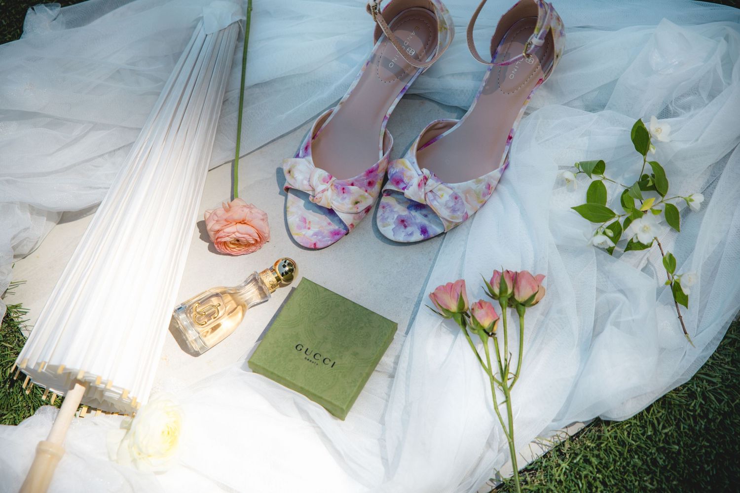 Floral wedding heels, perfume, green clutch, pink roses, and a white umbrella arranged on a bridal gown outdoors.