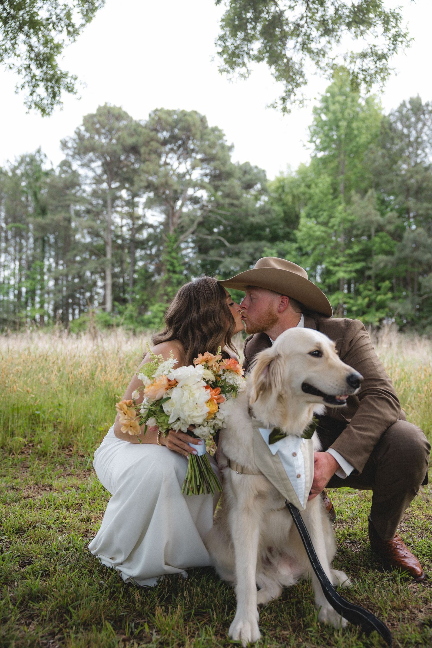 Bride and groom kissing outdoors with their golden retriever dog, bride holding floral bouquet, groom wearing cowboy hat.